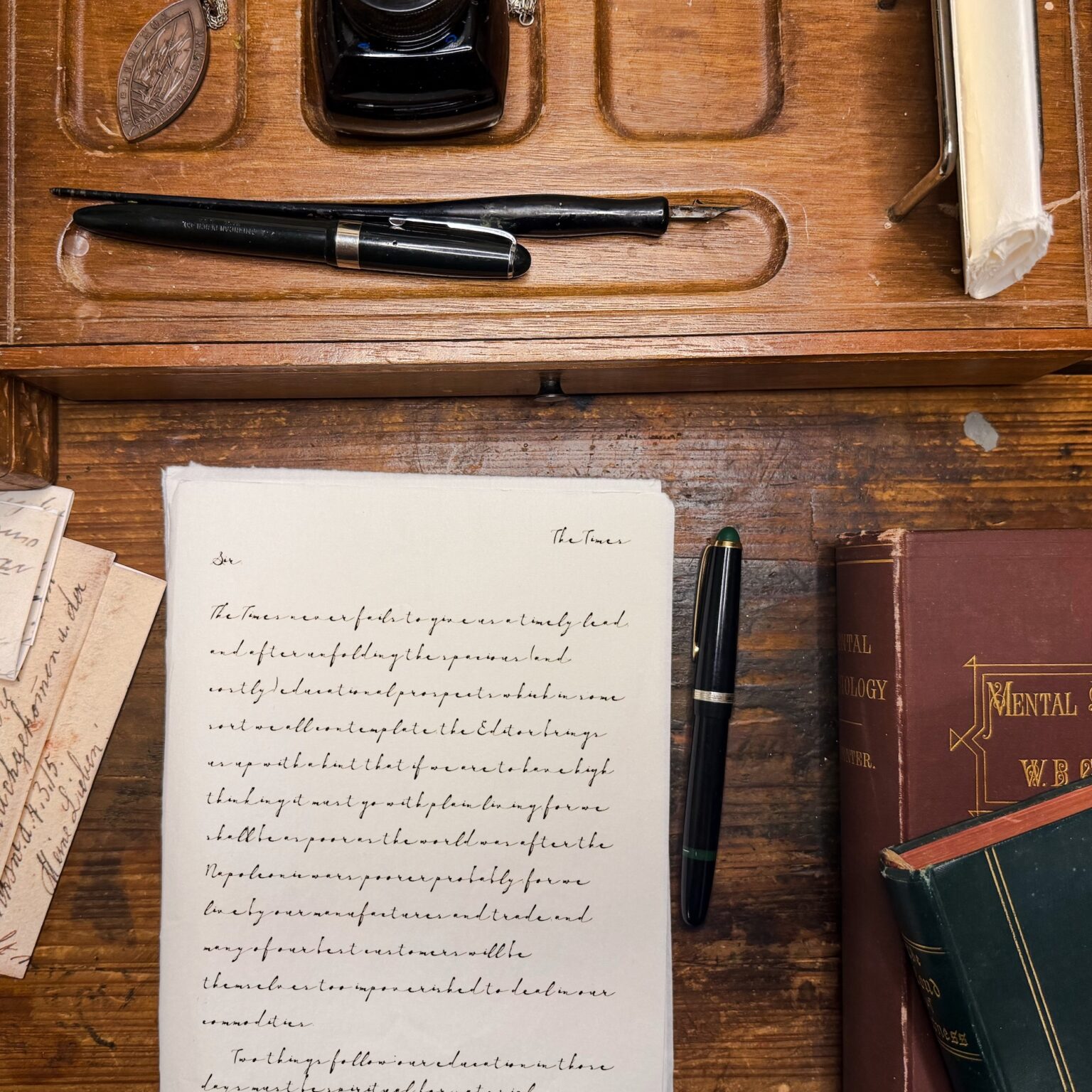Vintage desk scene with two black fountain pens laid over handwritten papers and a large book on a wooden surface for a writing setup image.