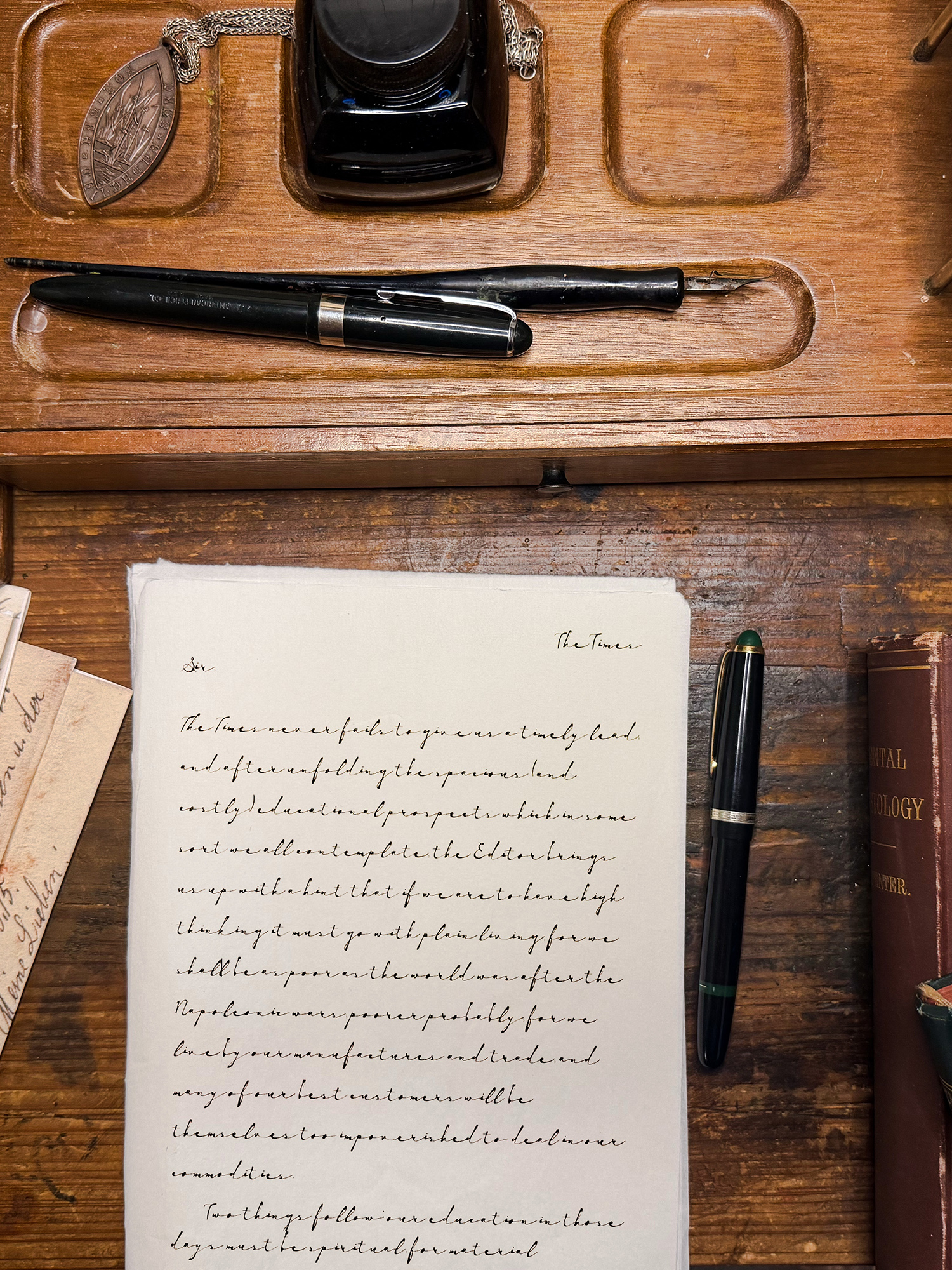 Antique writing desk scene with a black fountain pen, ink bottle, and a metal medallion on a chain beside a handwritten page.