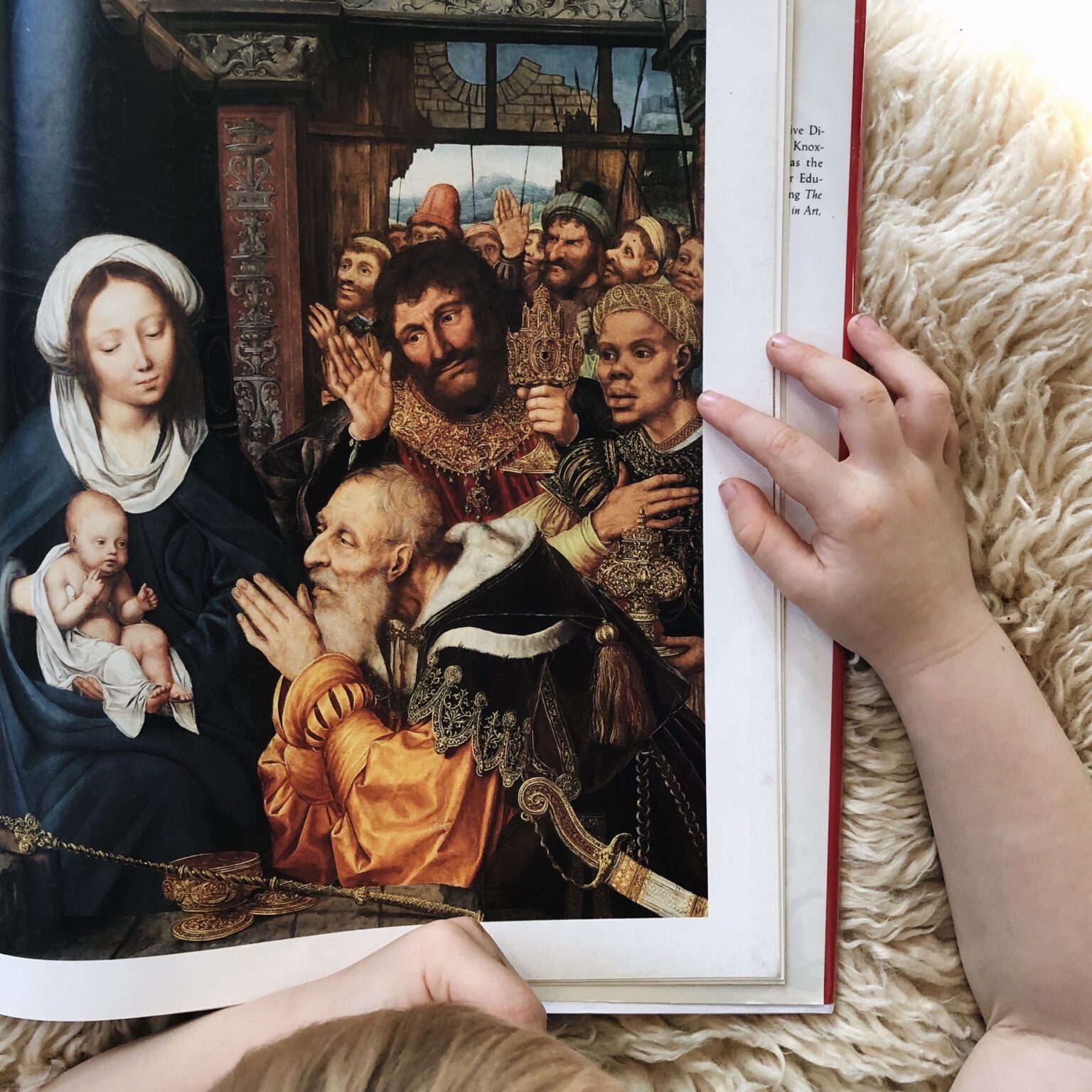 Open book page showing a detailed Renaissance scene: a bearded man in ornate robes blessing others, with a Madonna and child on the left; hands hold the page.