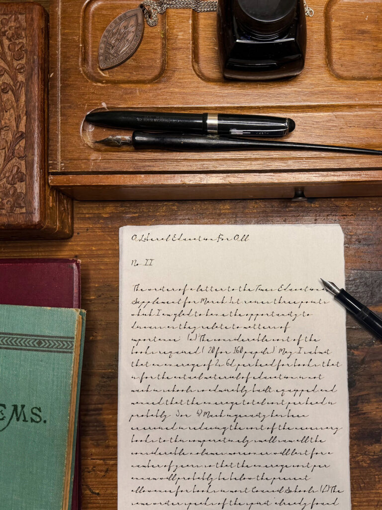 Open notebook with handwritten cursive page on a wooden desk, a black fountain pen, and an ink bottle nearby.