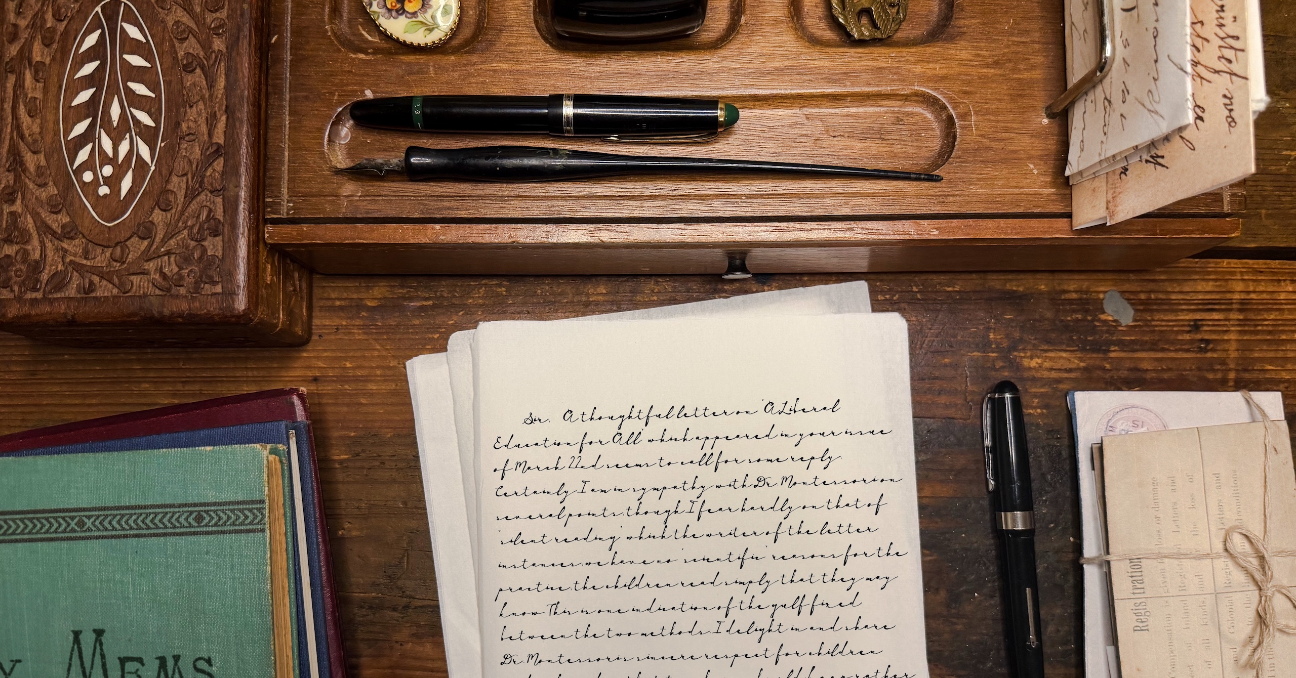 Vintage wooden desk with a black fountain pen, nib, and scattered handwritten letters beside worn books and a wooden drawer knob.