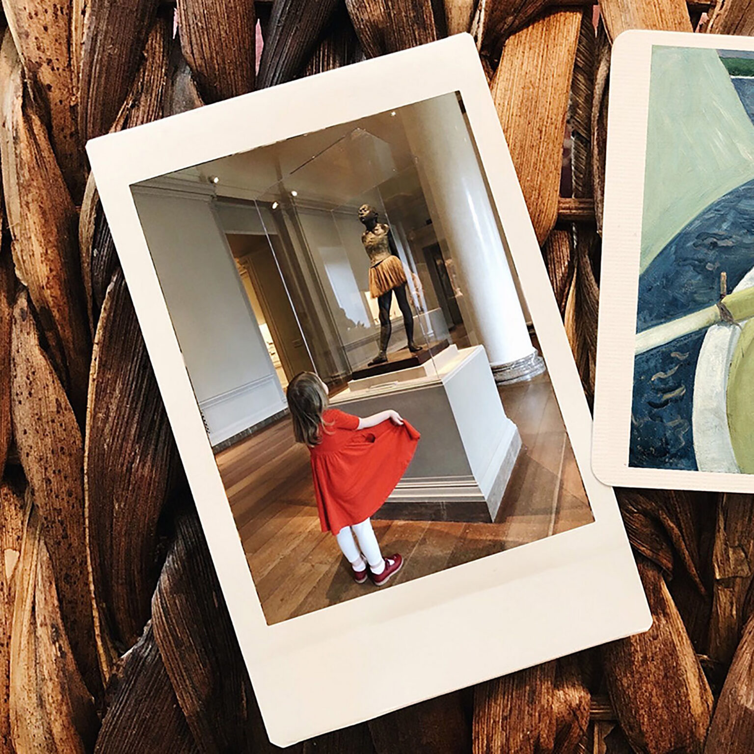Girl in a red dress stands inside a tilted white photo frame on a wooden floor/gallery backdrop.