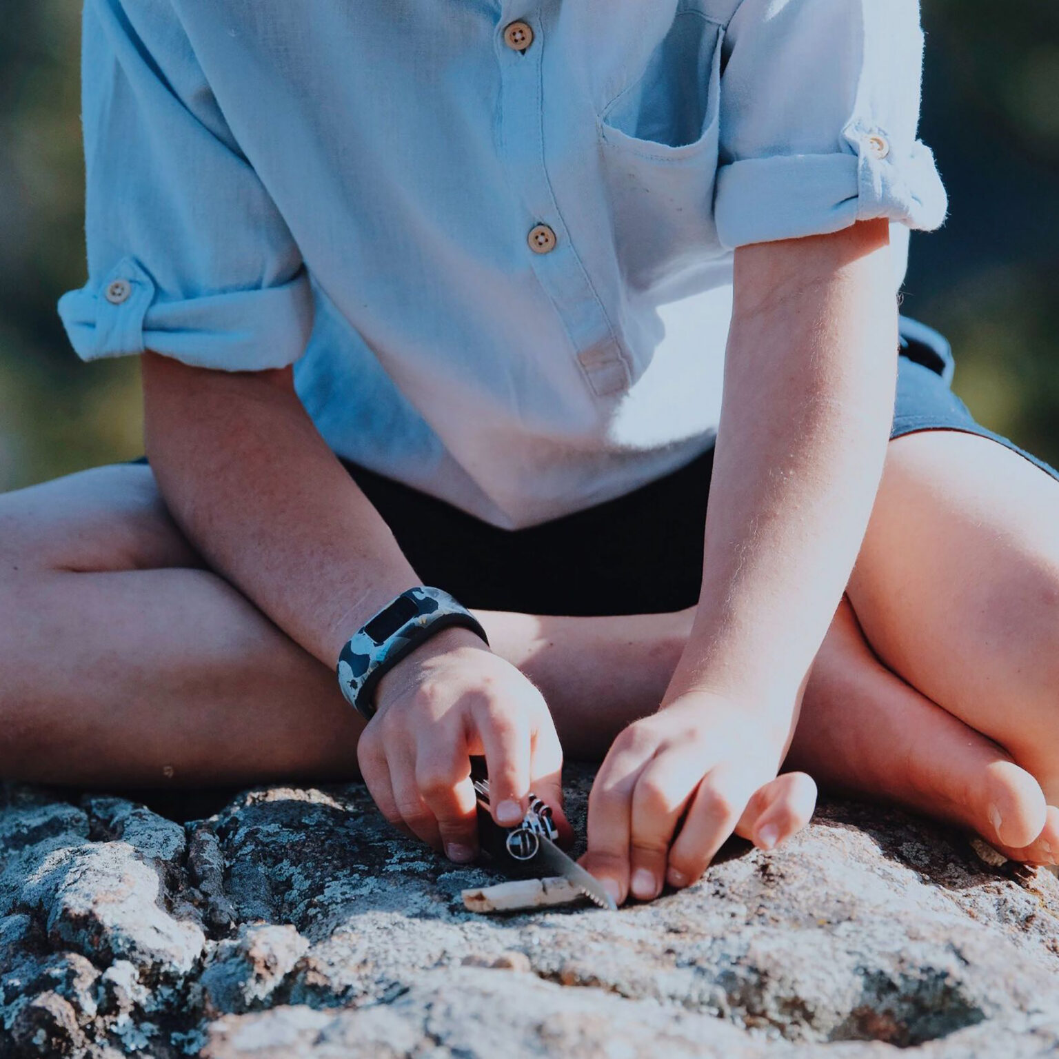Child wearing a light blue shirt, sleeves rolled up, playing with a small toy car on a rocky outdoor surface.