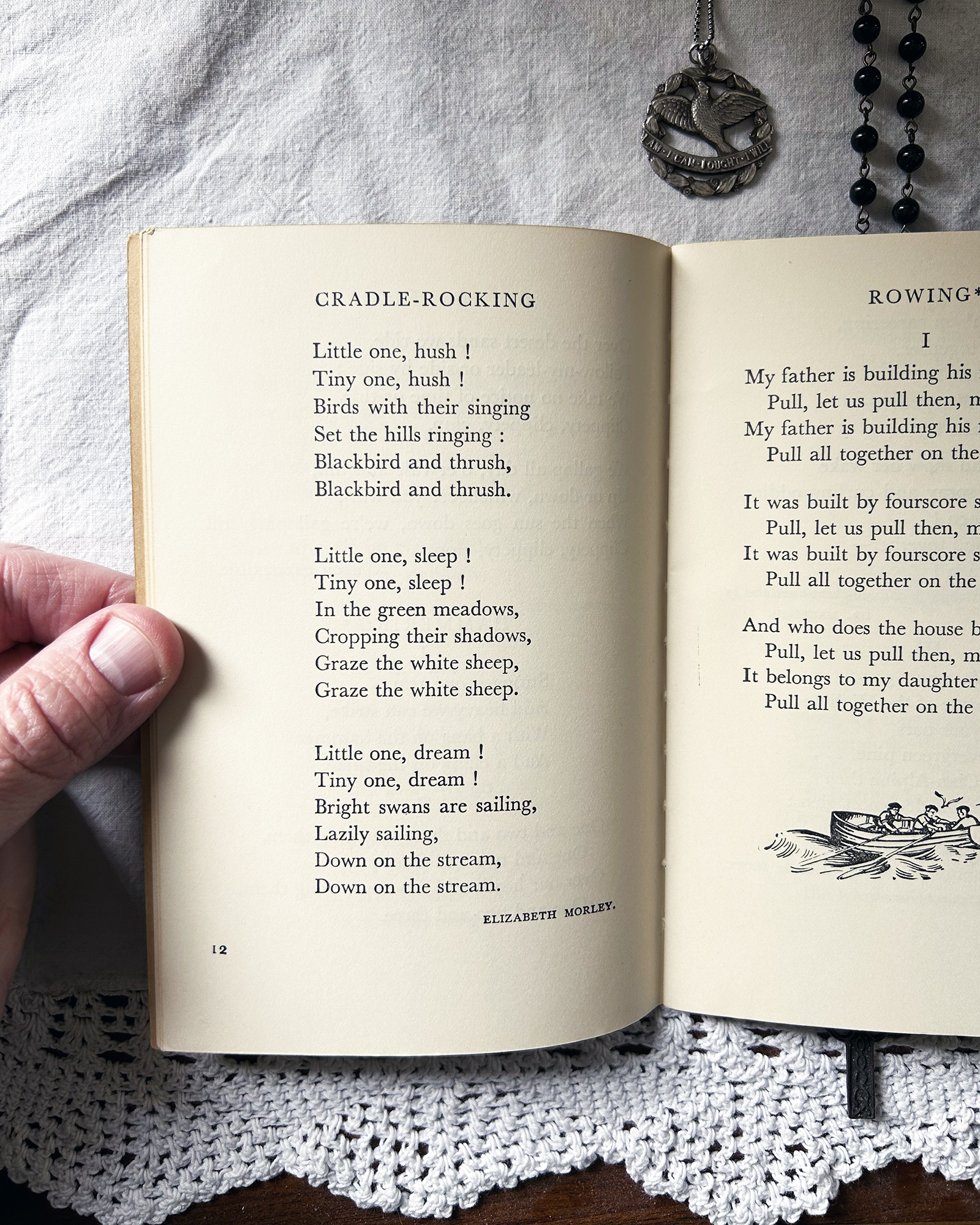 Open book with the poem 'Cradle-Rocking' visible on the left page, hand at the bottom left, and decorative jewelry at the top right over a lace doily surface.