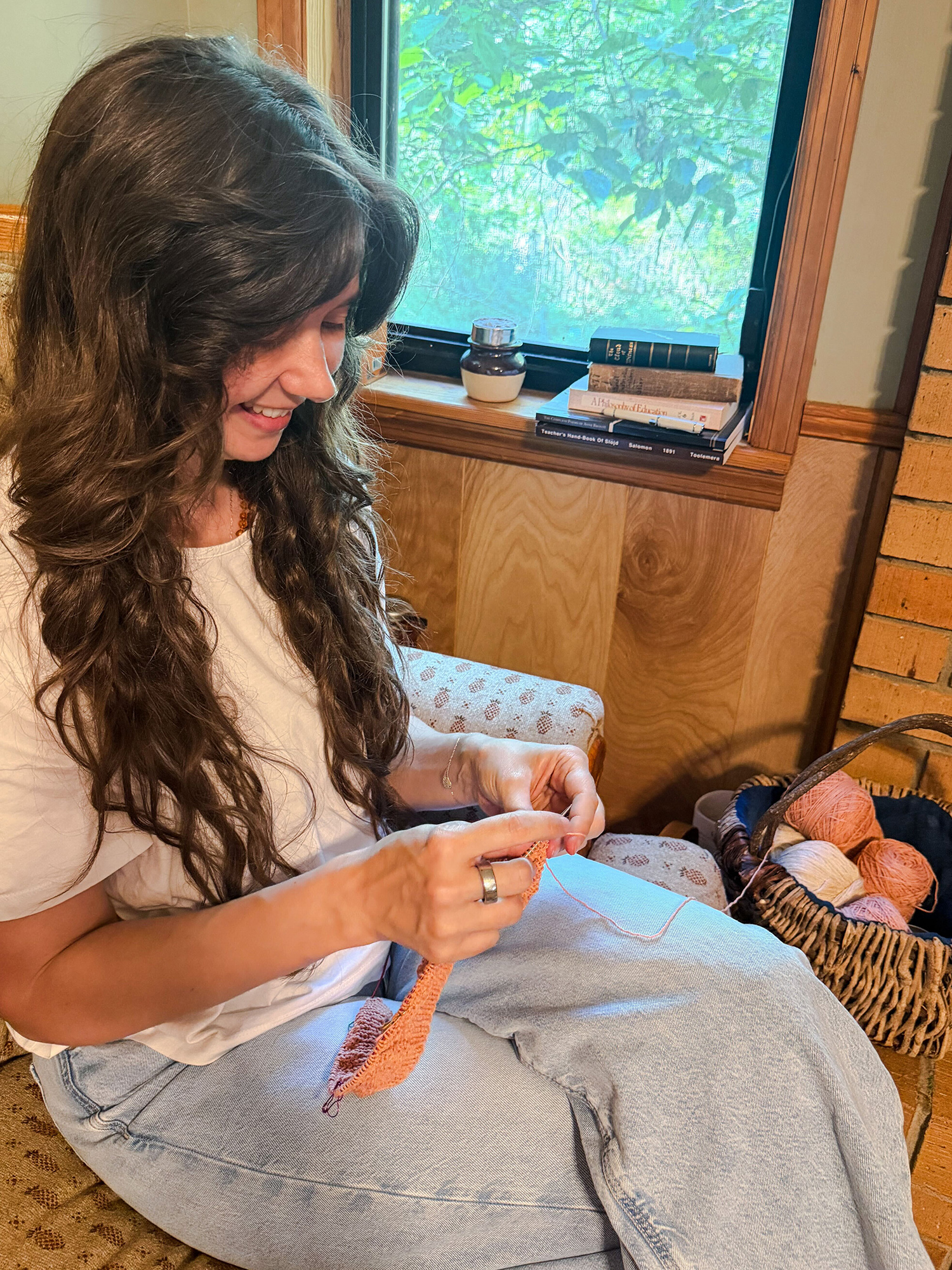 Woman with long dark hair knitting orange yarn on her lap by a window.