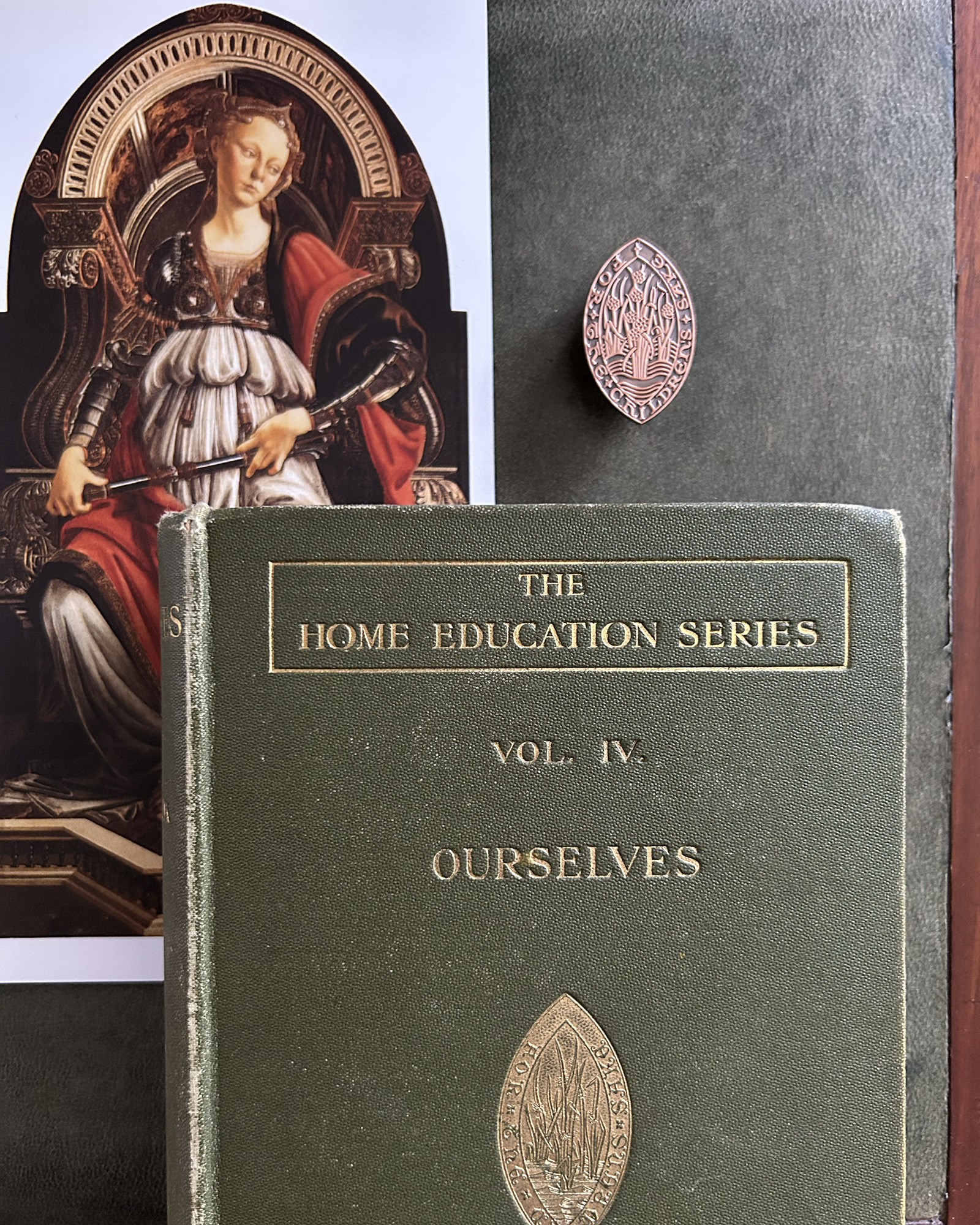 Portrait of a woman in armor seated on a throne on the left, with a red cape; to the right, a small oval engraved metal pin on a dark book cover reading 'The Home Education Series, Vol. IV, Ourselves'