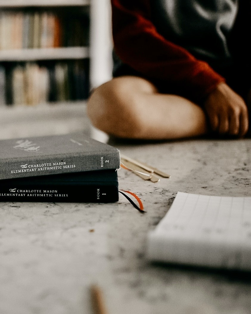 Close-up of arithmetic books and study materials on a desk, with a person sitting cross-legged in the background.