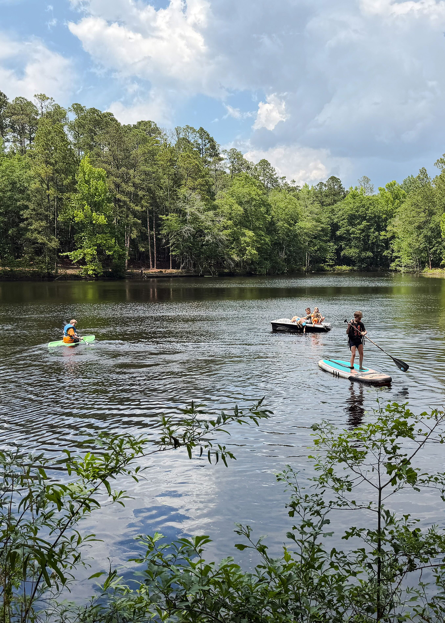 People enjoying a lakeside scene: a green kayak with a paddler, a small motorboat with riders, and a paddleboarder on calm water, all beside a tree-lined shore under a partly cloudy sky.