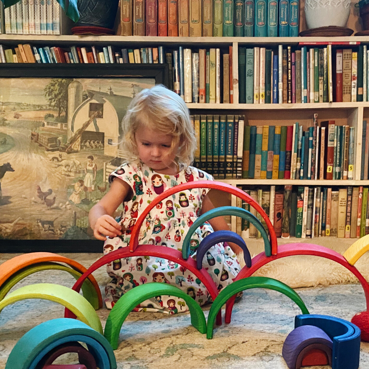 Child in a patterned dress playing with colorful circular rings in a library or classroom setting, with tall bookshelves in the background and a vintage poster nearby.
