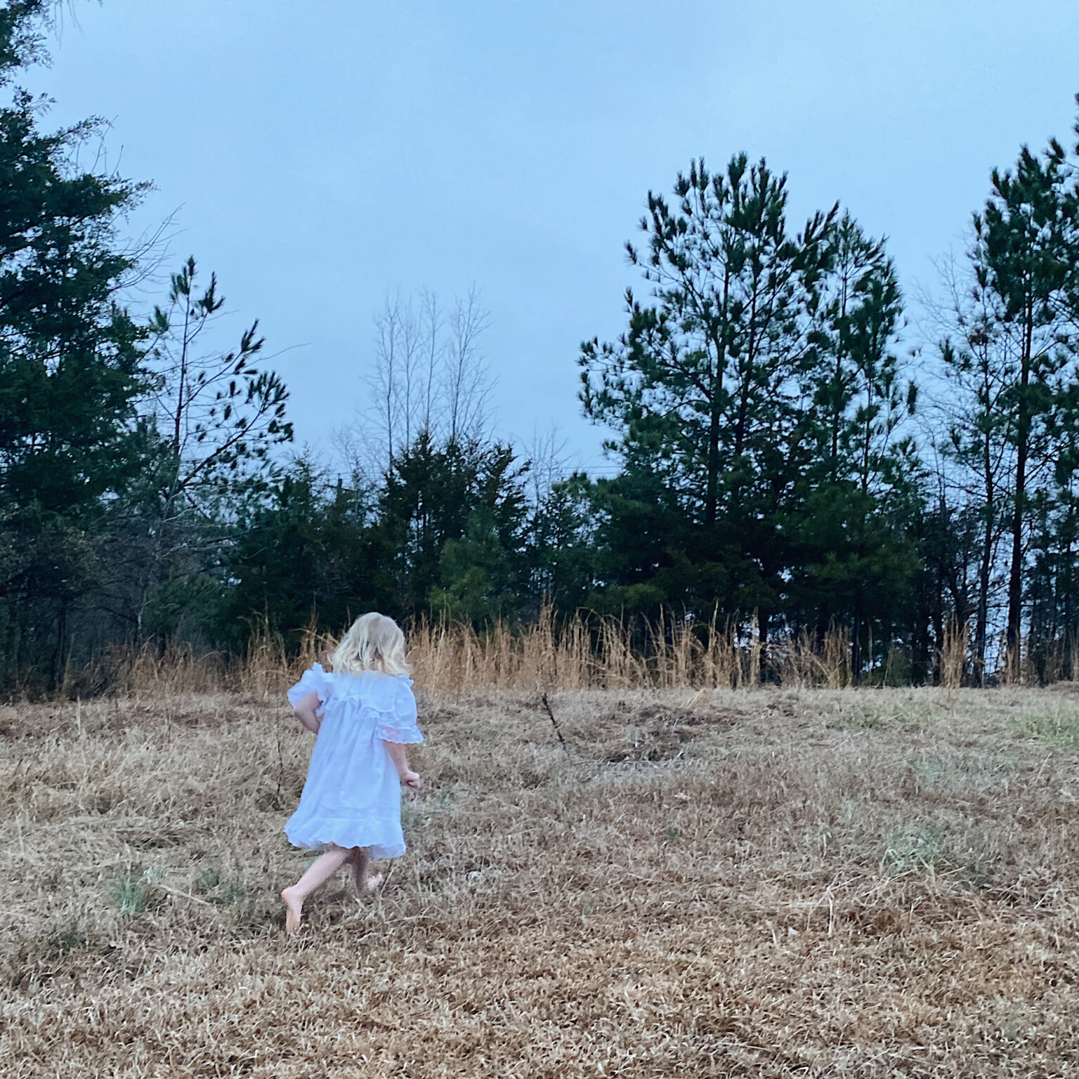 A young child in a white dress runs barefoot across a dry grassy field with evergreen trees in the background.