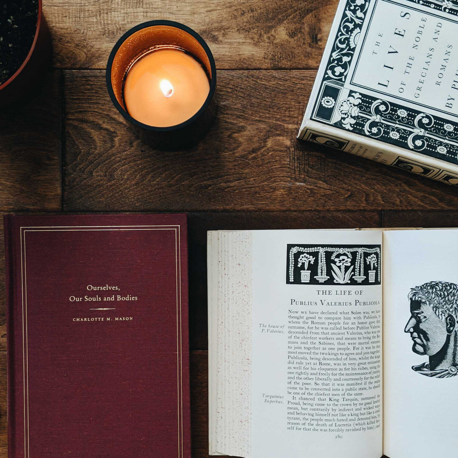 Lit candle in a black holder on a wooden table beside an open antique book with ornate illustrations and a burgundy cover nearby.