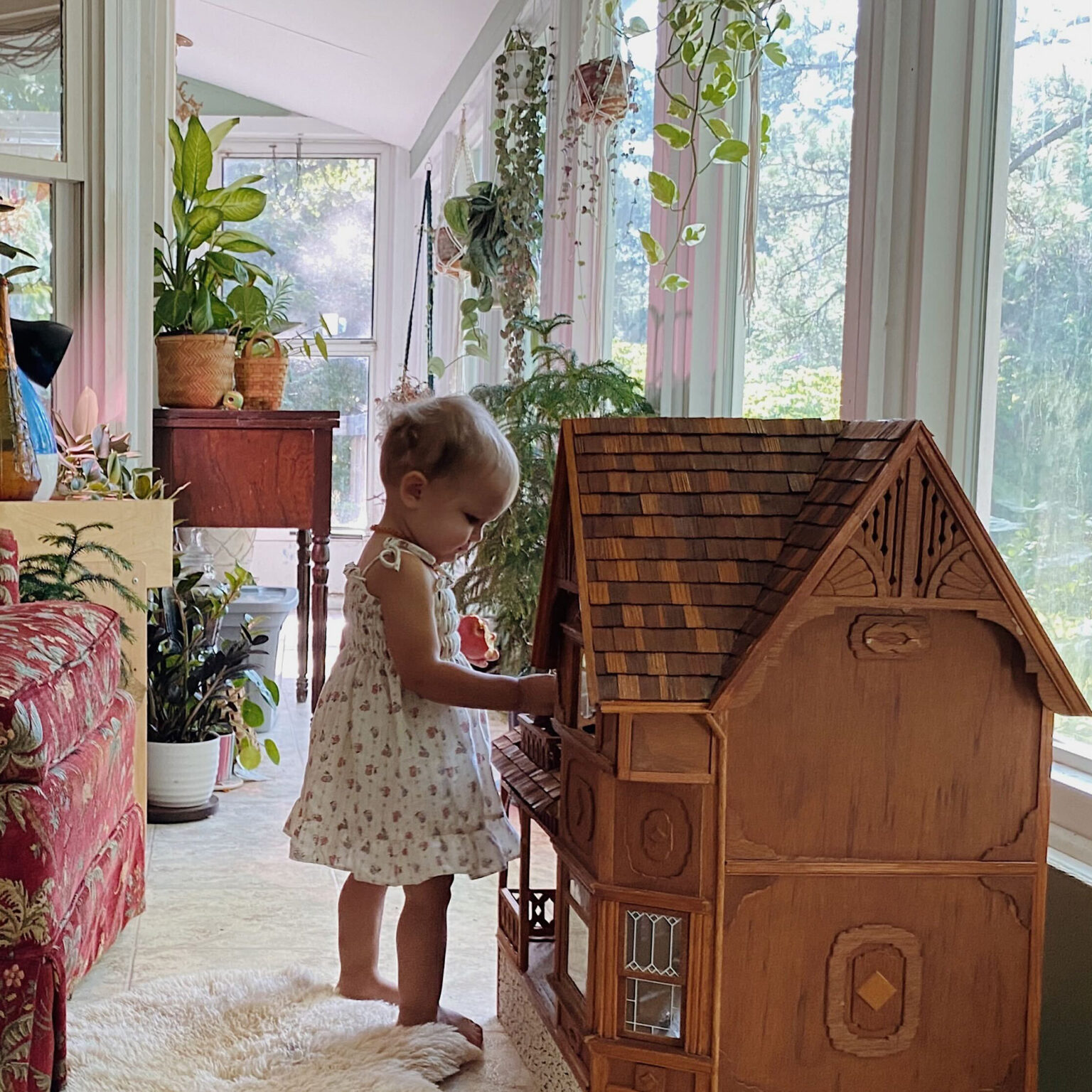 A young girl in a floral dress stands barefoot and plays a large wooden toy piano in a sunlit, plant-filled room with windows.
