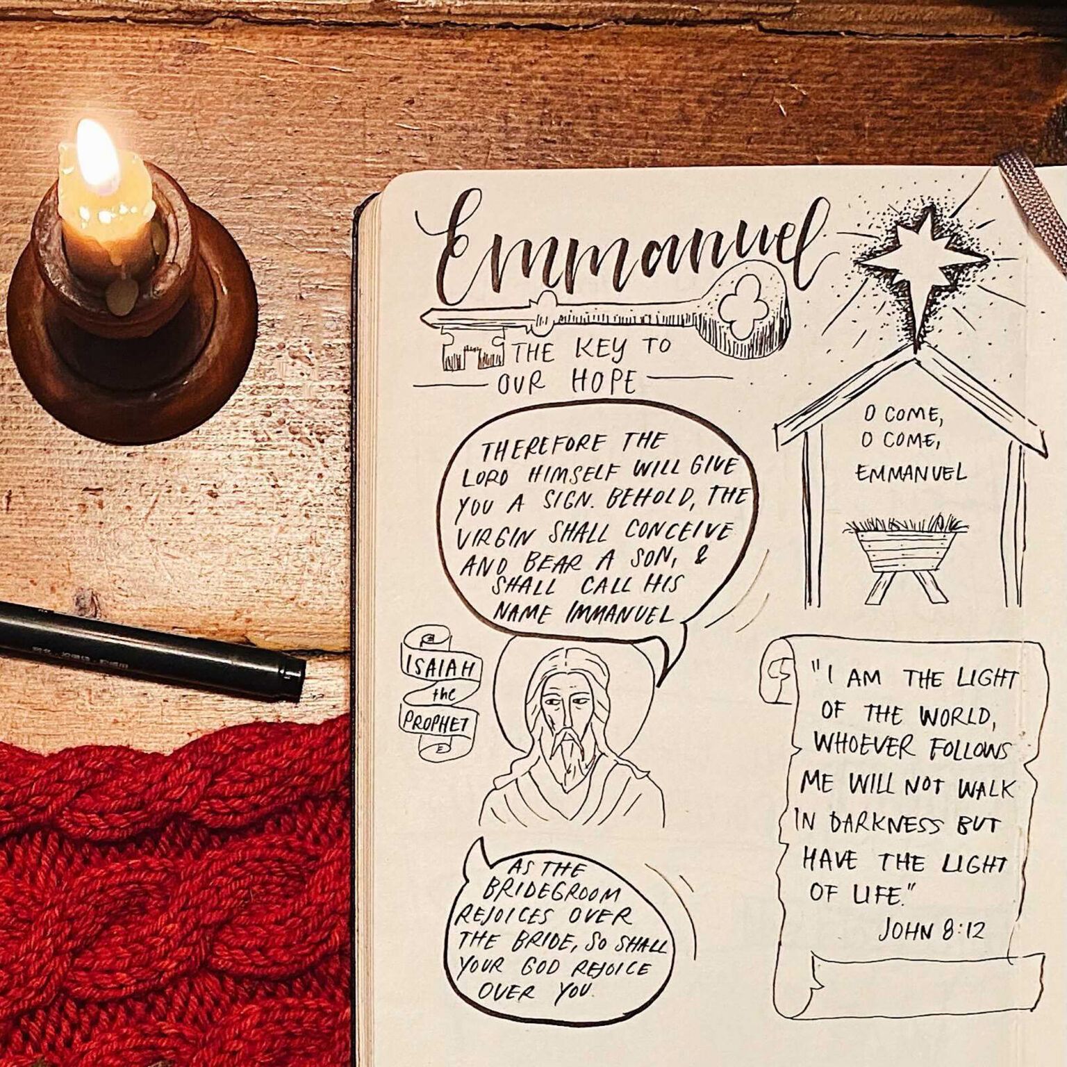 Open notebook page with hand-drawn biblical illustration and the word 'Emmanuel,' beside a lit candle on a wooden table.
