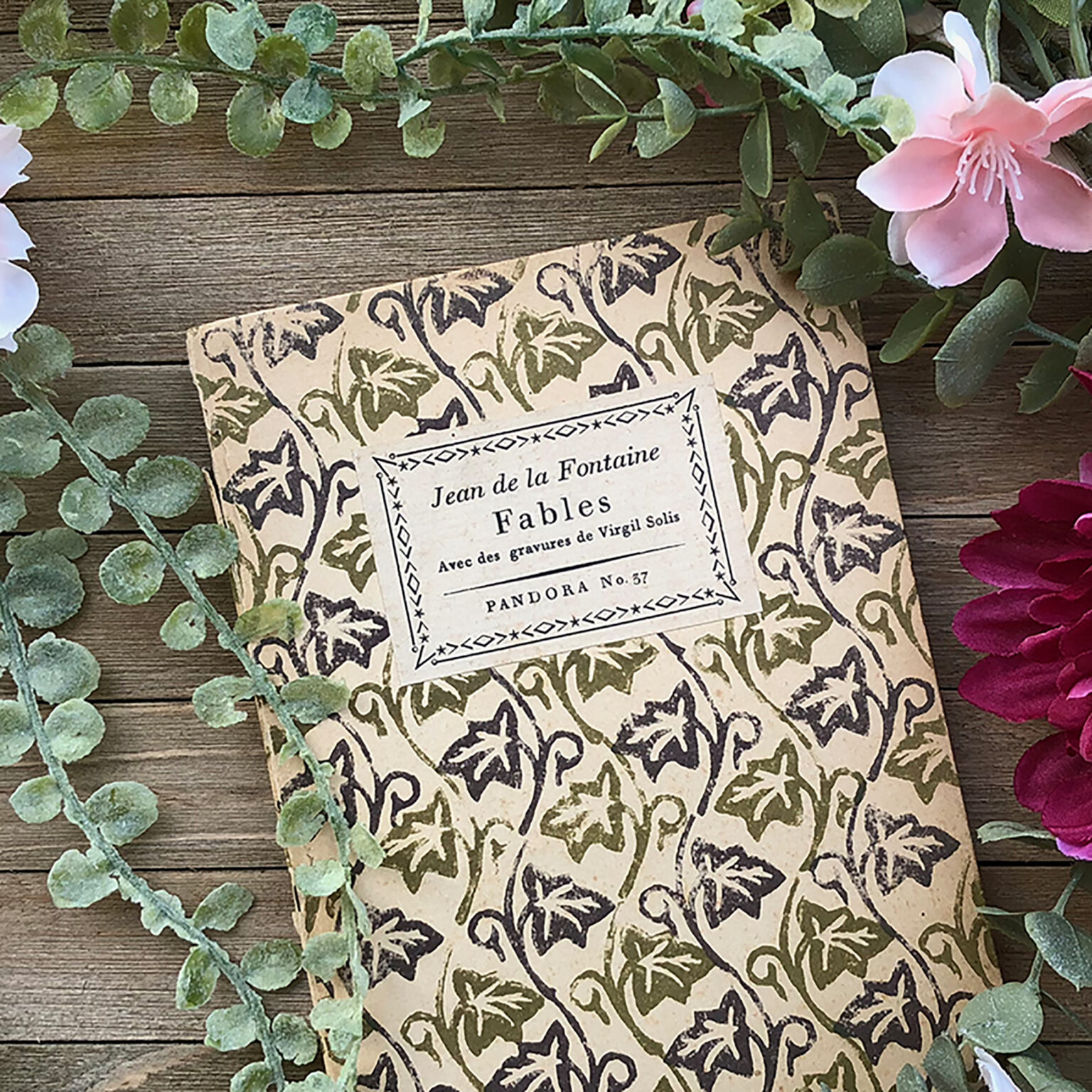 Flat lay of vintage book pages with a decorative black border, surrounded by green leaves and pink petals on a wooden table surface.125 chars