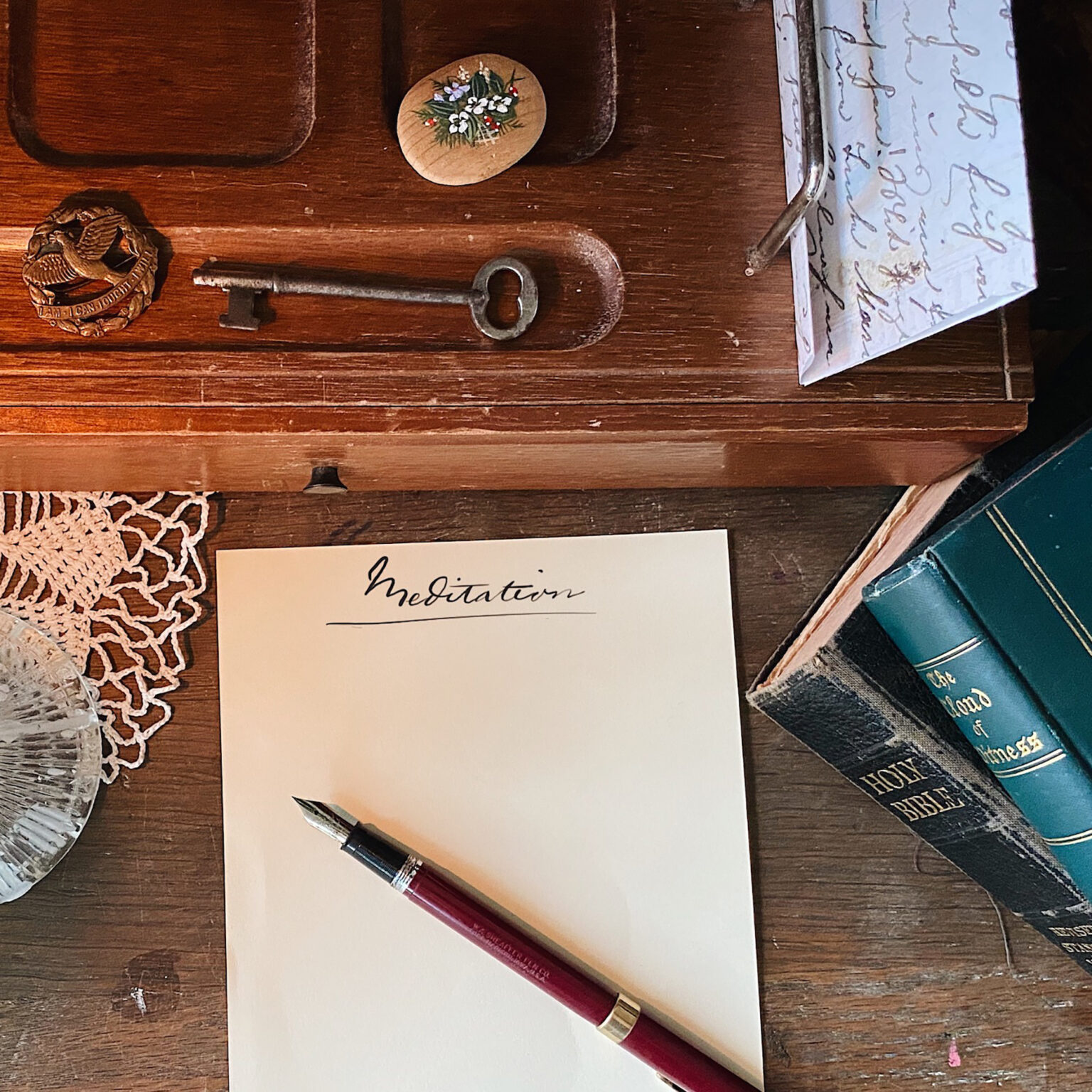 Top-down view of a wooden desk with a red fountain pen on a blank card, an antique key, and a floral-painted stone.