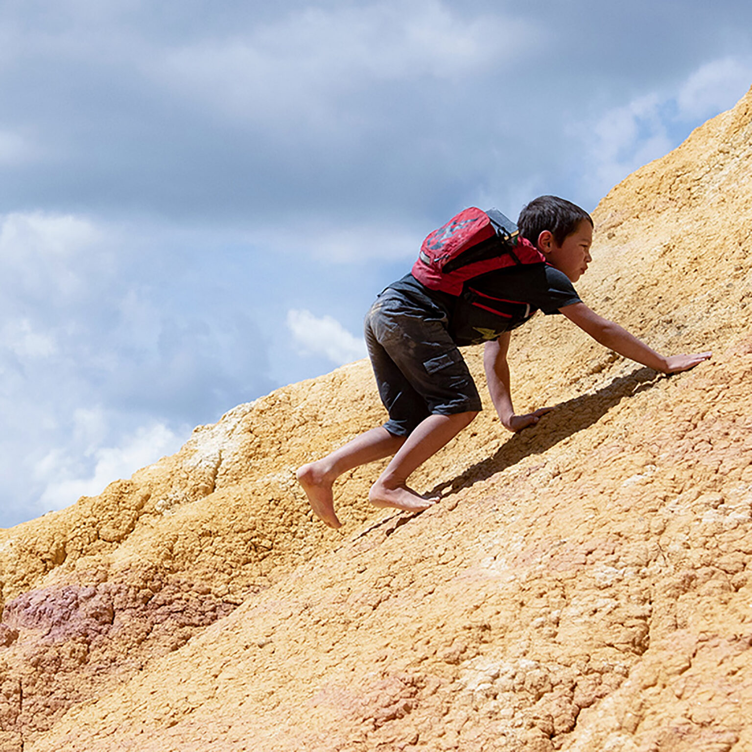 Boy with a red backpack climbs up a sandy yellow hillside under a blue sky outdoors.
