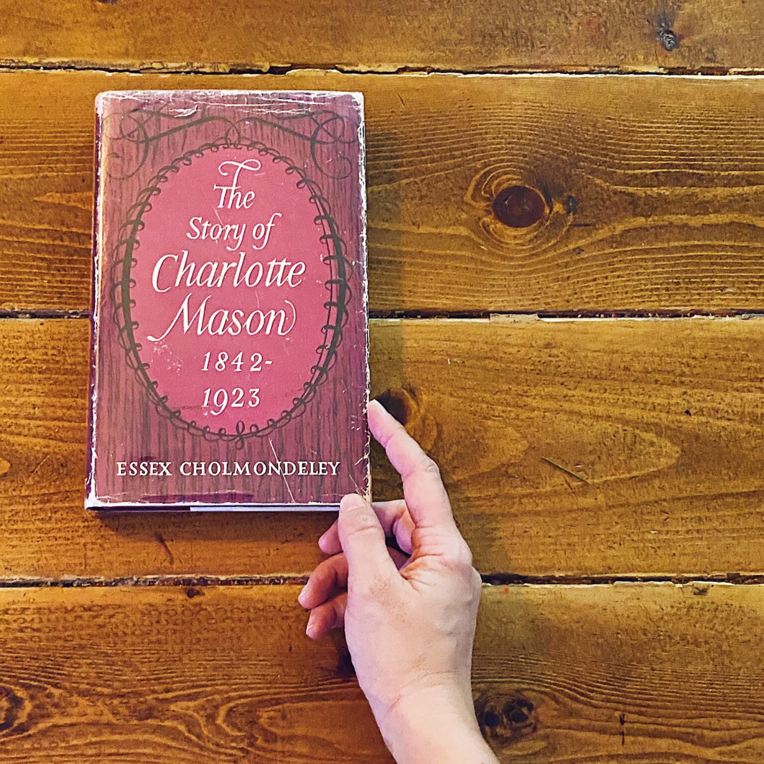 Pink hardcover book titled 'The Story of Charlotte Mason' held by a hand on a wooden table surface.