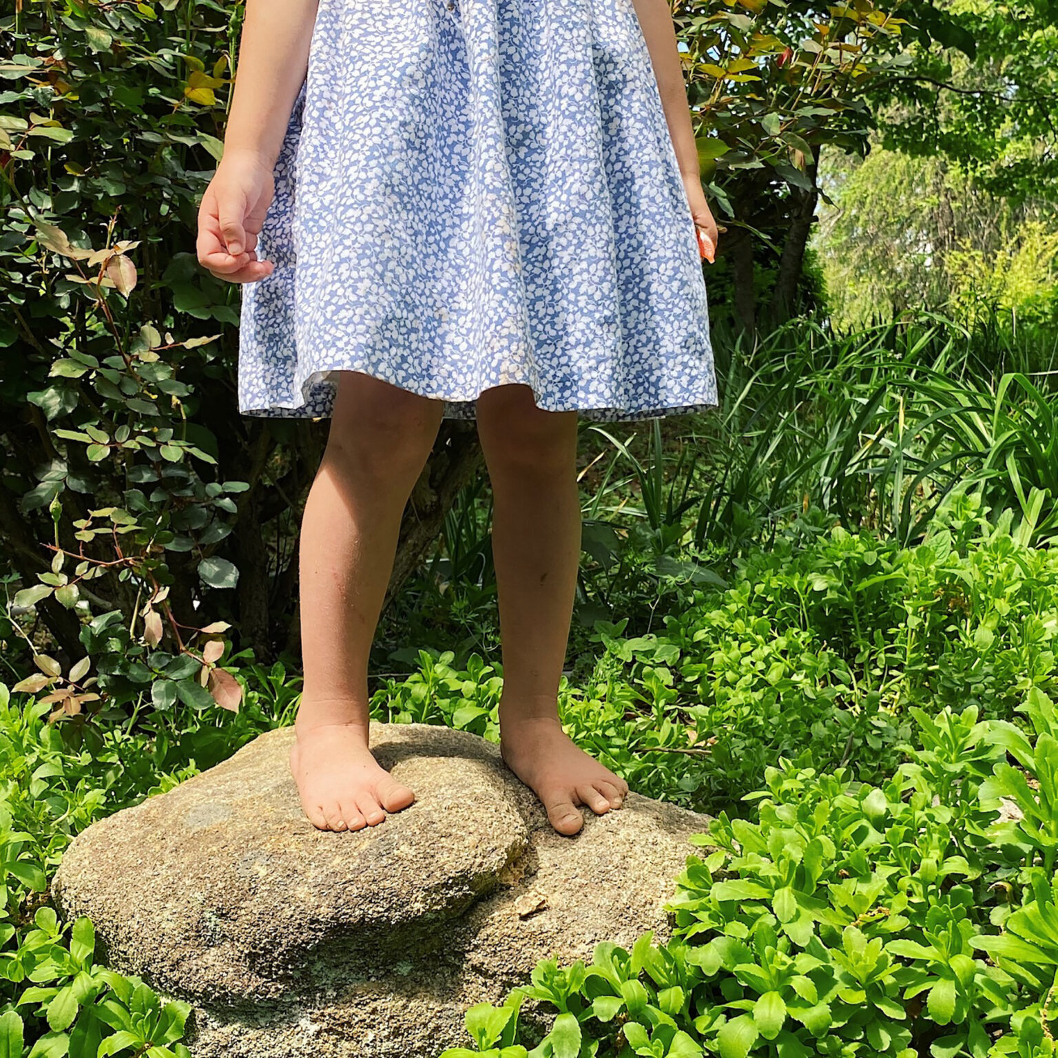 Child standing barefoot on a rock in a garden, wearing a light blue floral dress.
