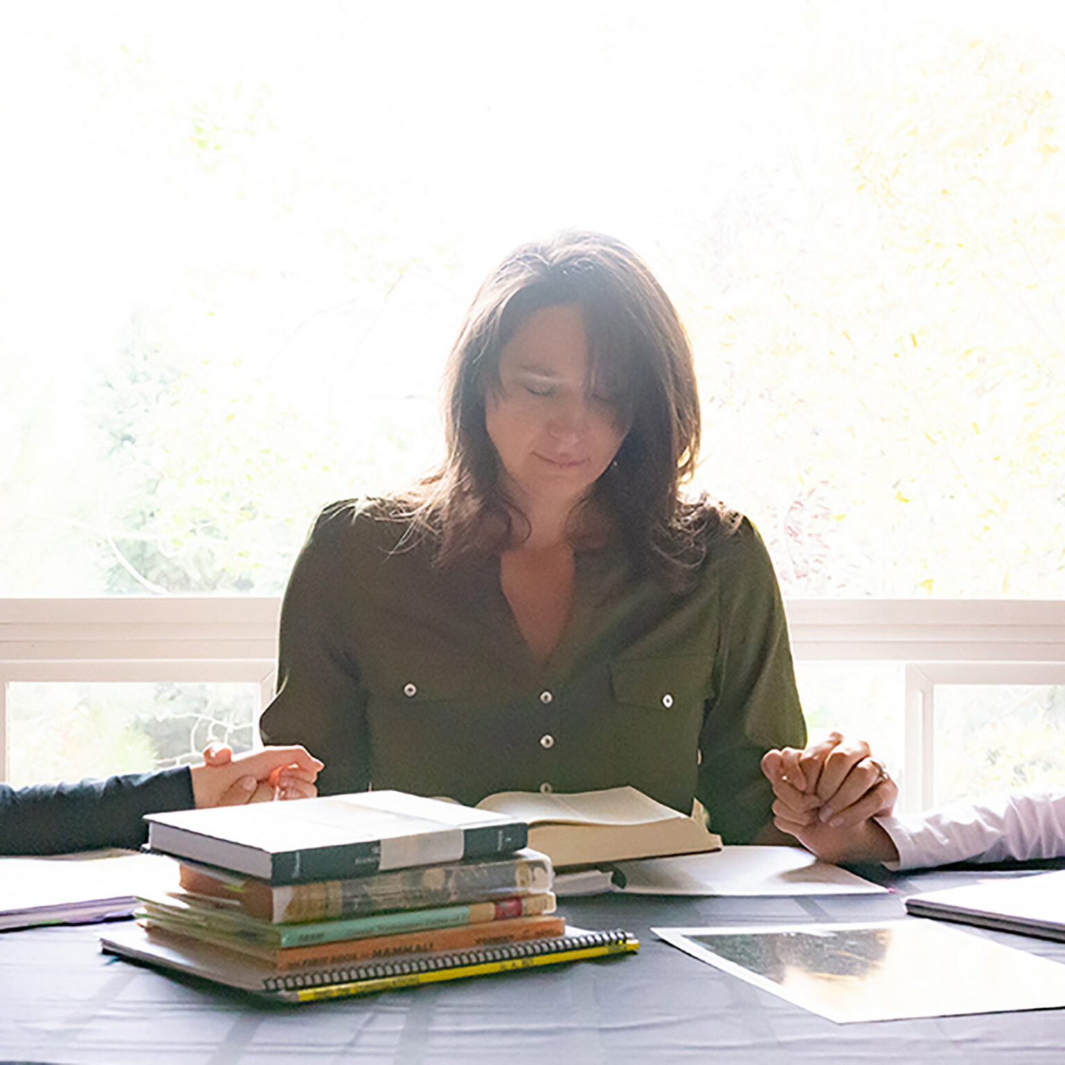 Woman in a dark green blouse sits at a table with stacked notebooks and loose papers, another person’s hands visible at the edges of the frame.