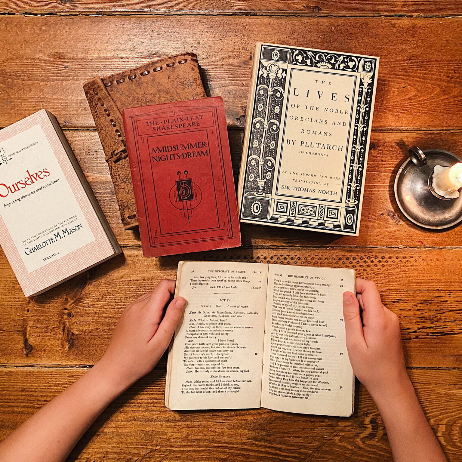 Two hands hold open an old book with small print on a wooden table, flanked by other vintage books and a metal candle holder in warm light.