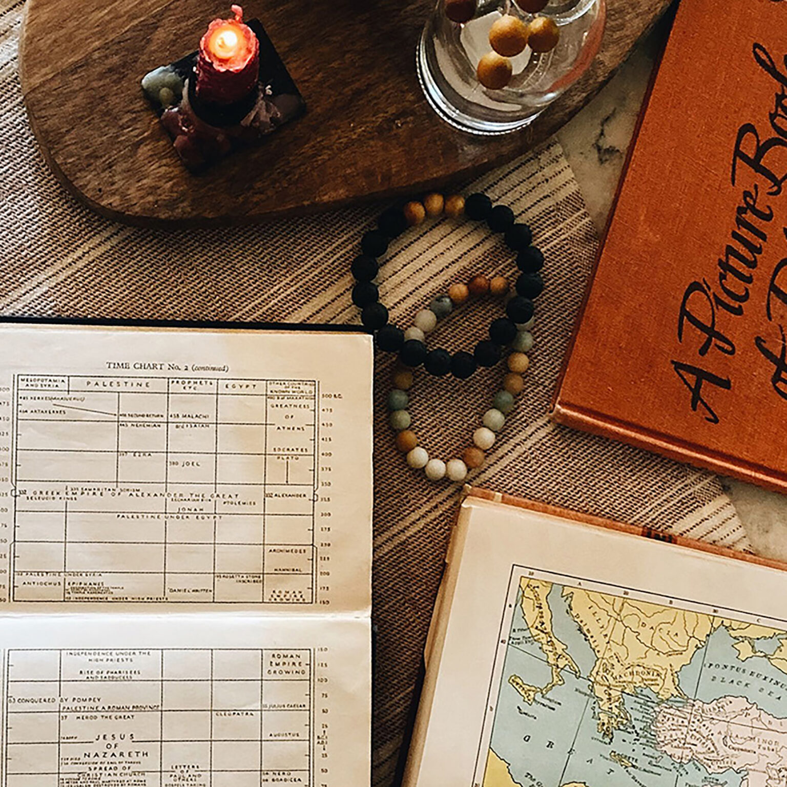 Lit red candle, beaded necklace, old documents, and a map on a textured cloth—vintage still life.