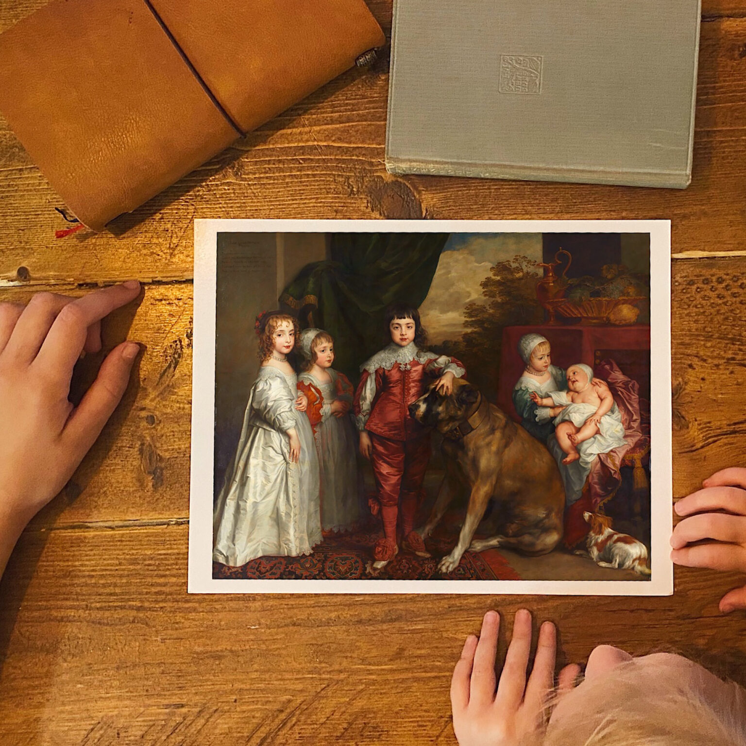 Overhead shot of a wooden table with a printed painting, a tan leather journal, and a gray book, with several hands around the edges.