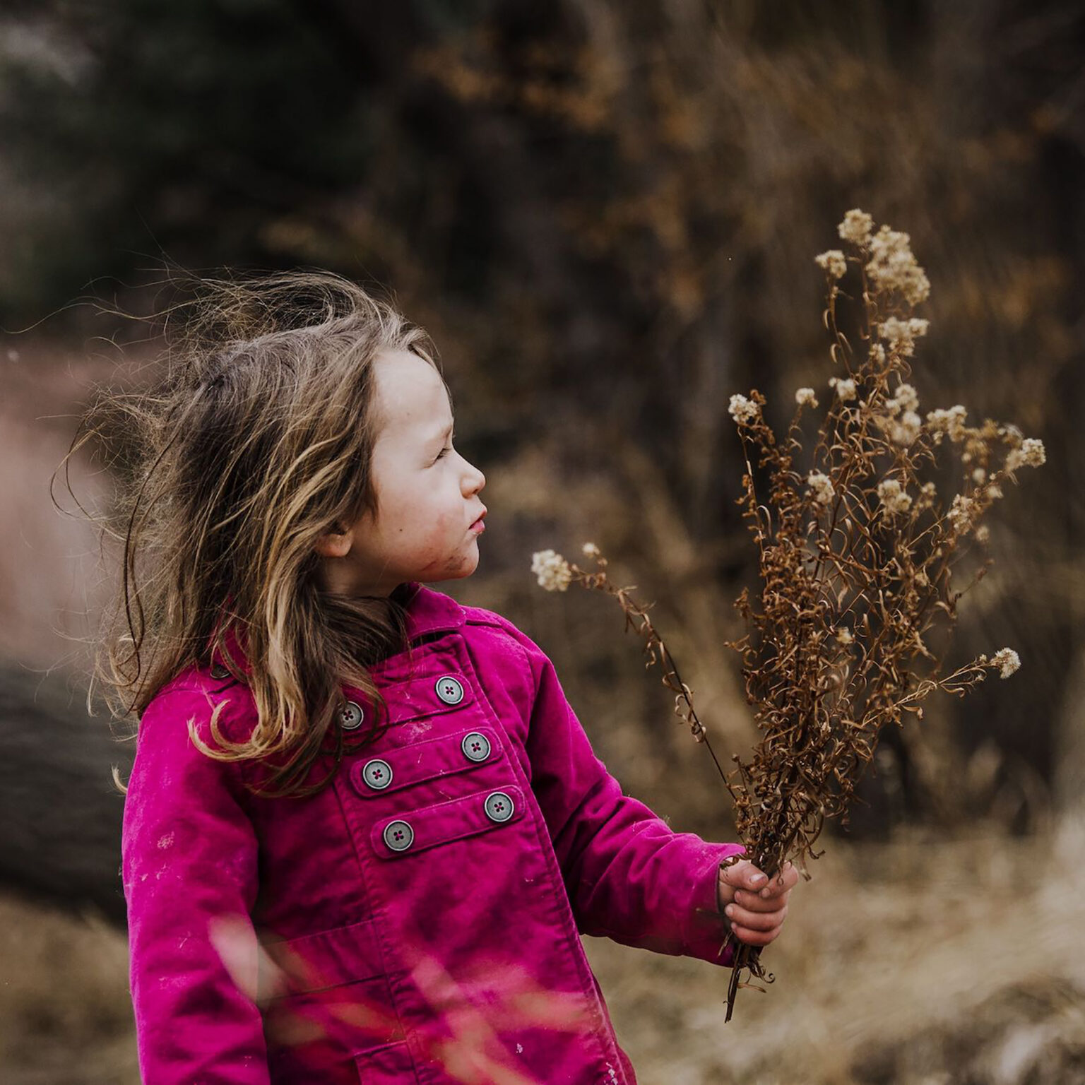 Young girl in a bright pink jacket stands outdoors, softly holding a dried plant.