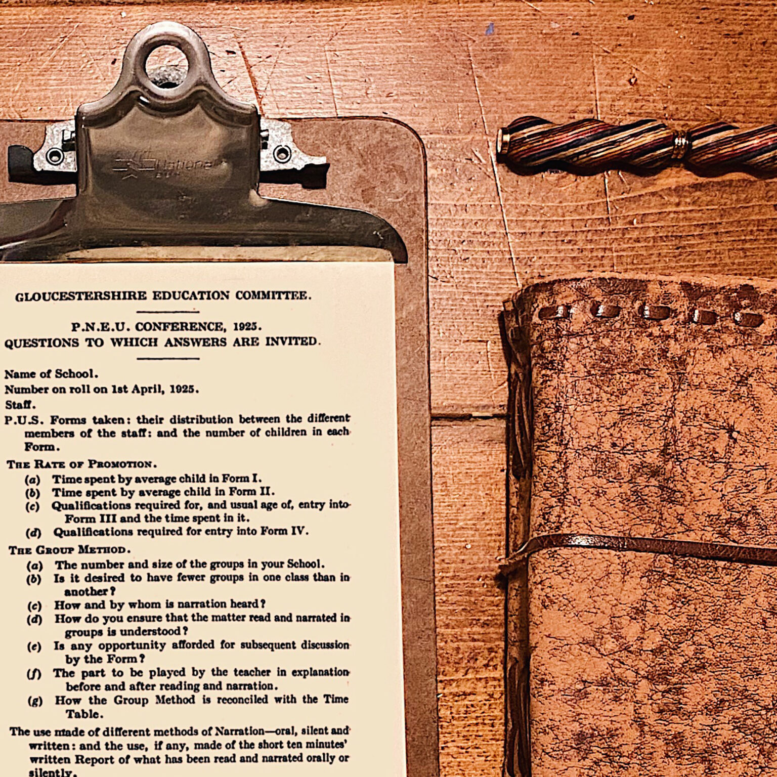 Close-up of a vintage metal clipboard holding a 1925 Gloucestershire Education Committee document on a wooden table. The carved wooden handle is visible on the right edge.