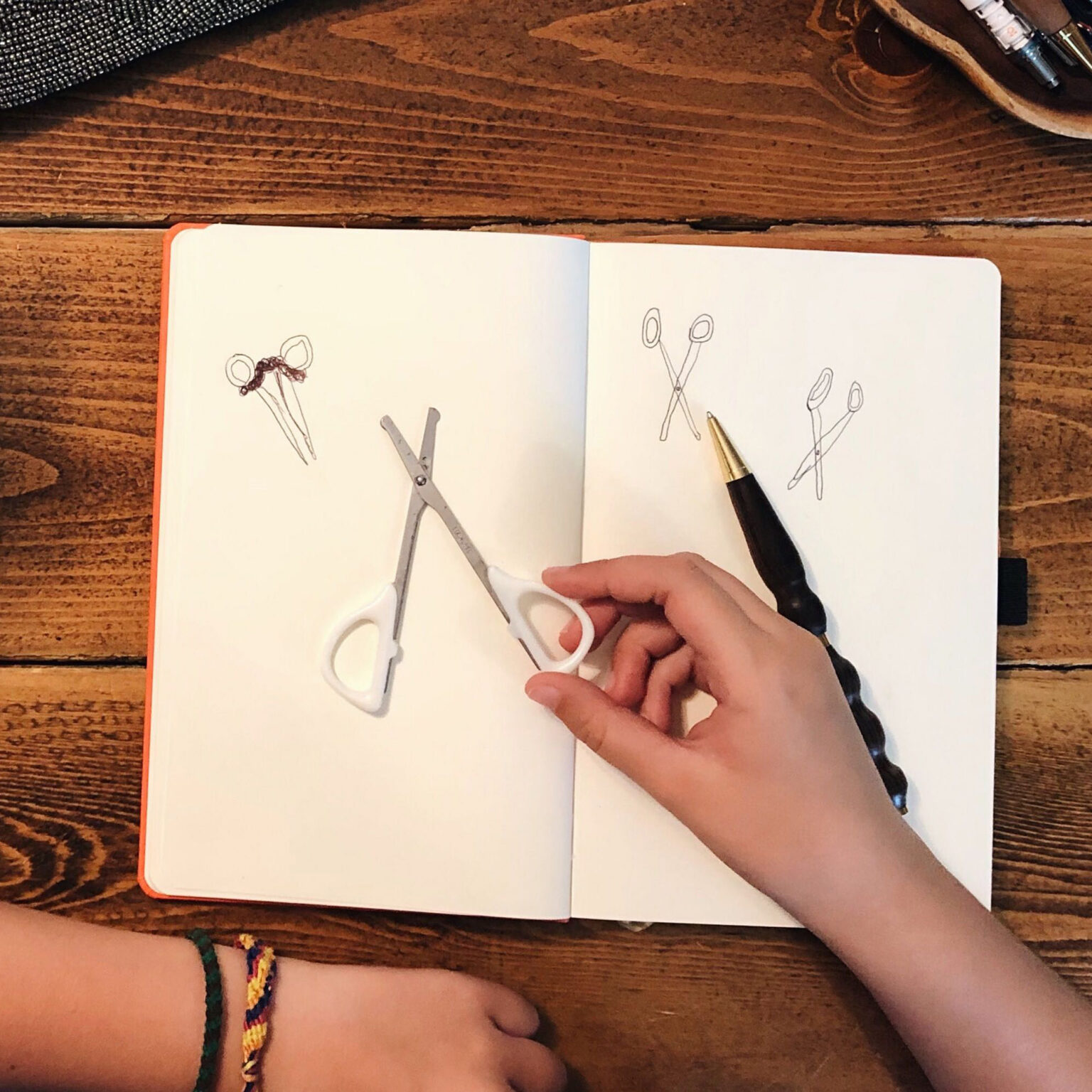 Open notebook on a wooden table with sketches of scissors; a person holds white-handled scissors and a craft knife nearby, while a textured fabric swatch and markers sit at the edges.
