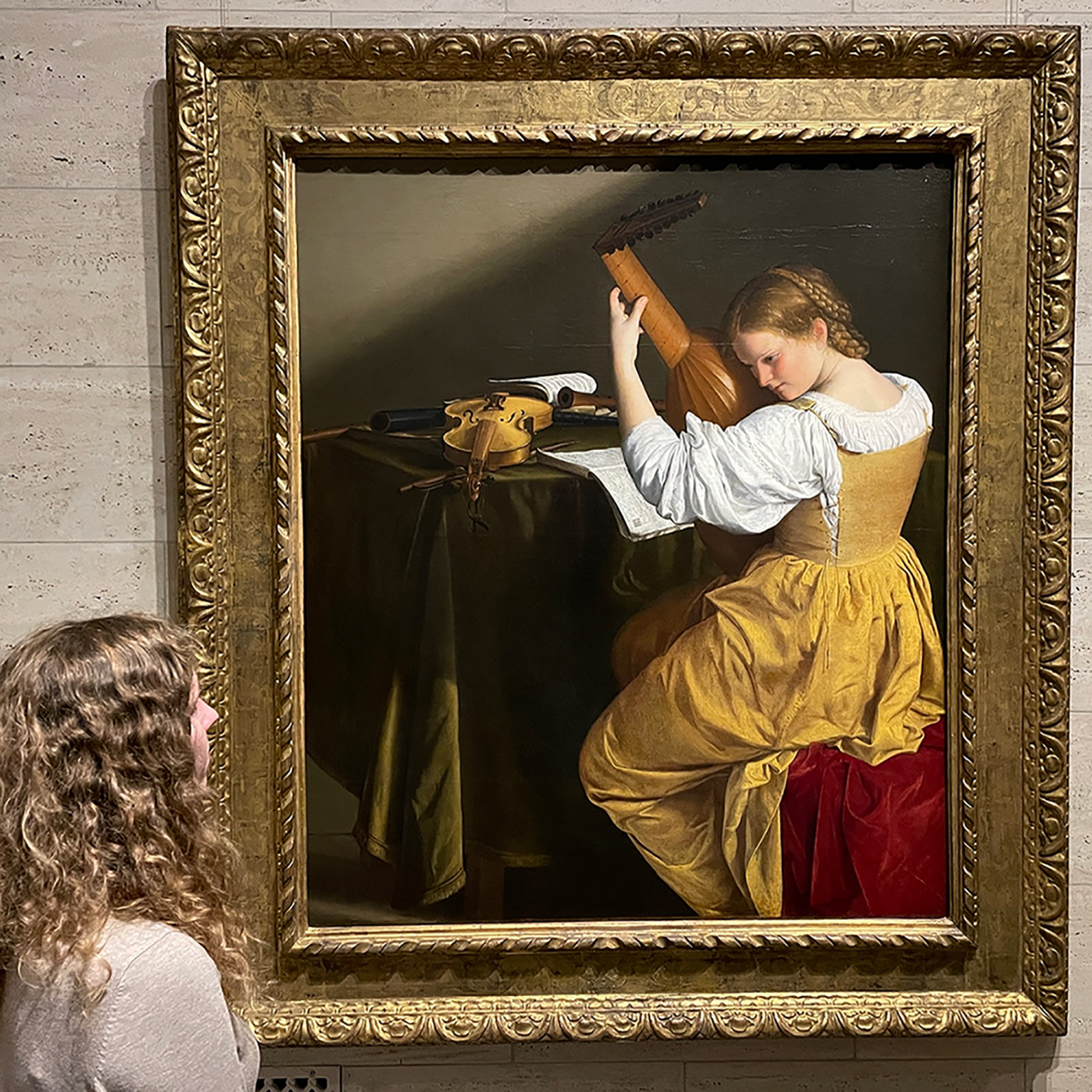 Framed classical painting of a girl in a white dress playing a violin, inside an ornate gold frame in a gallery setting.