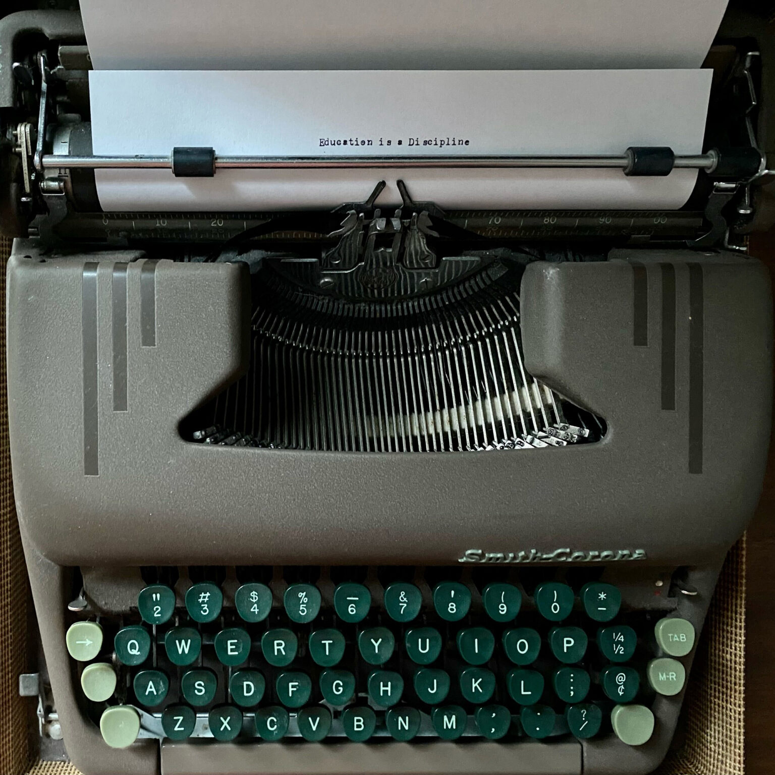 Vintage Smith-Corona typewriter with teal circular keys and a sheet of paper that reads 'Education is a Discipline' in typewriter font, viewed from the front.