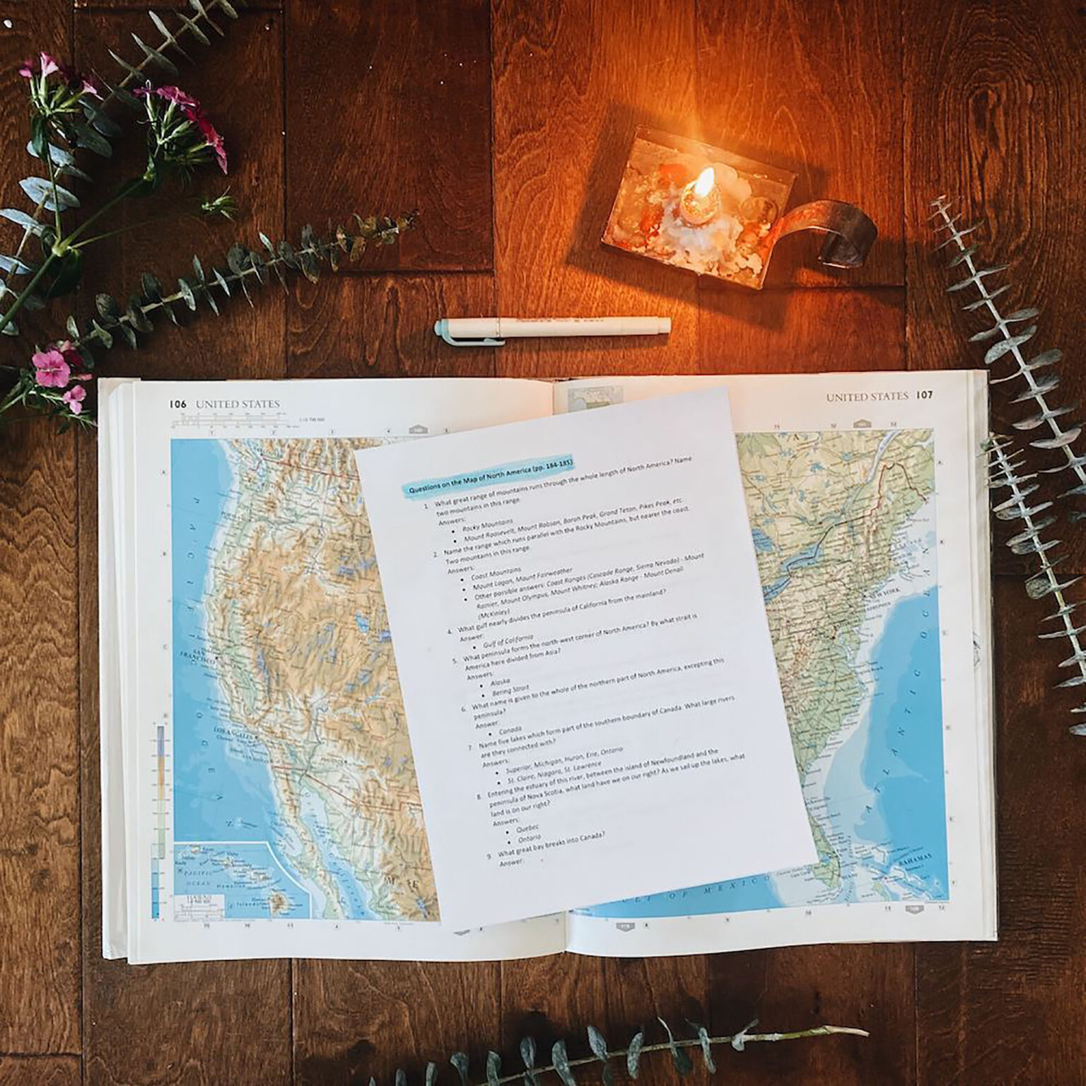 Overhead view of an open atlas on a wooden table with a lit candle and scattered papers and a pen nearby.