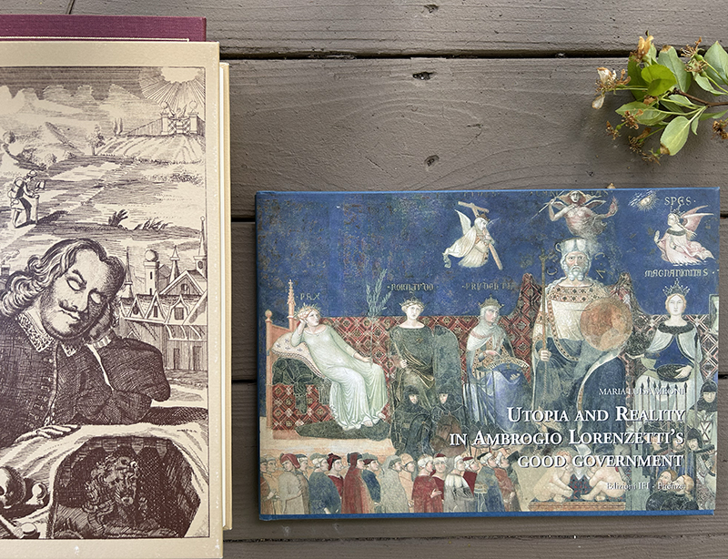 Two books on a wooden table: the left shows an illustrated page with a sleeping man, the right is a blue cover about Utopia and Reality in Ambrogio Lorenzetti’s Good Government, with a small plant in the top-right corner.