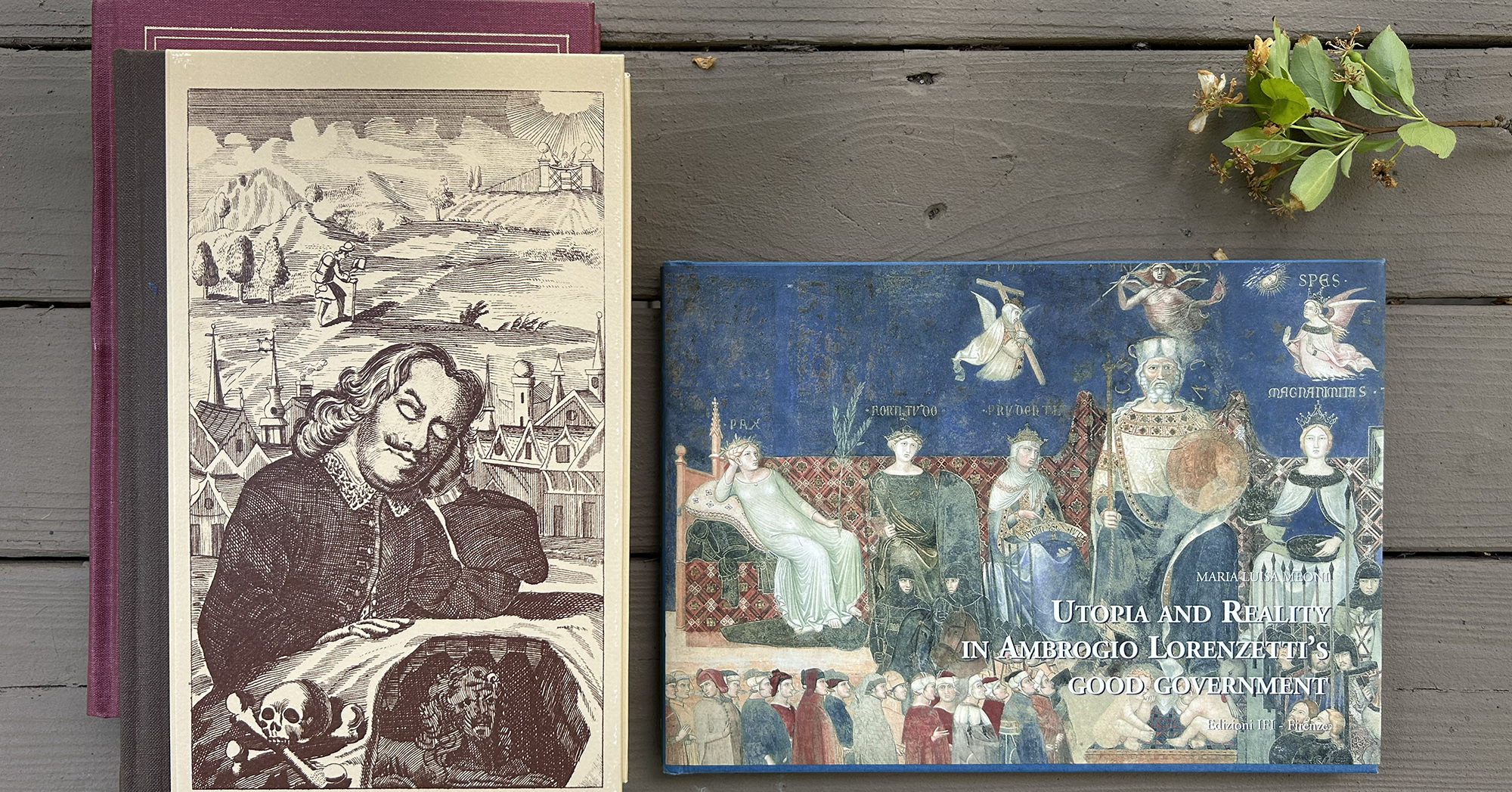 Two vintage books on a weathered wooden surface; a beige engraved landscape book beside a blue illustrated volume with a Renaissance scene and title text.