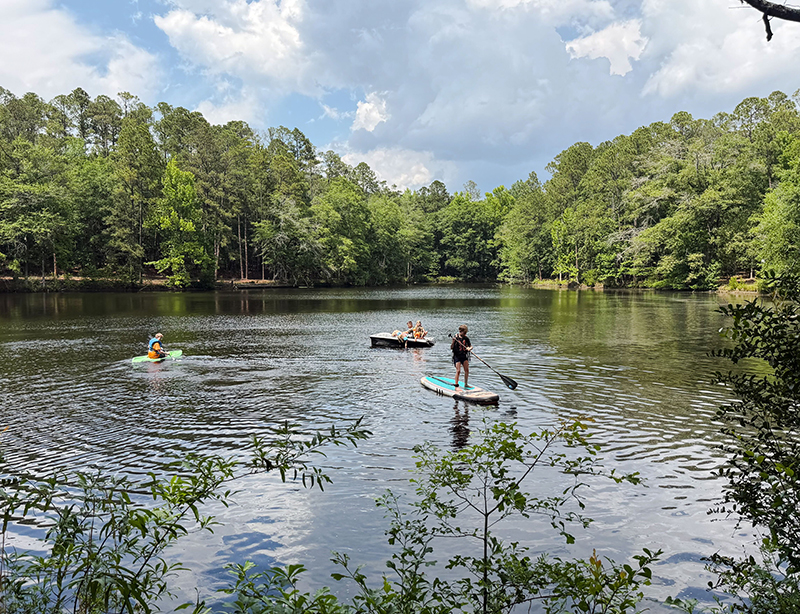 People paddleboarding on a calm lake with a tree-lined shore under a partly cloudy sky.