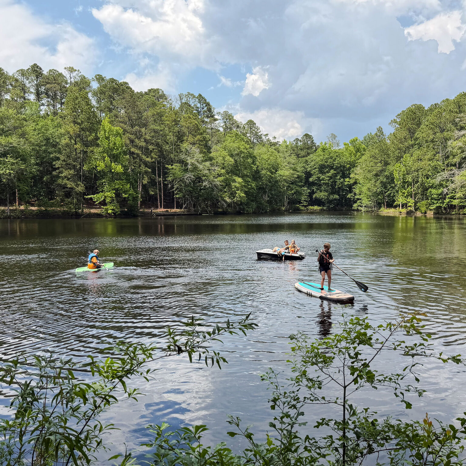 People in life jackets paddleboarding and kayaking on a calm lake with a tree-lined shore and a small boat nearby under a partly cloudy sky.