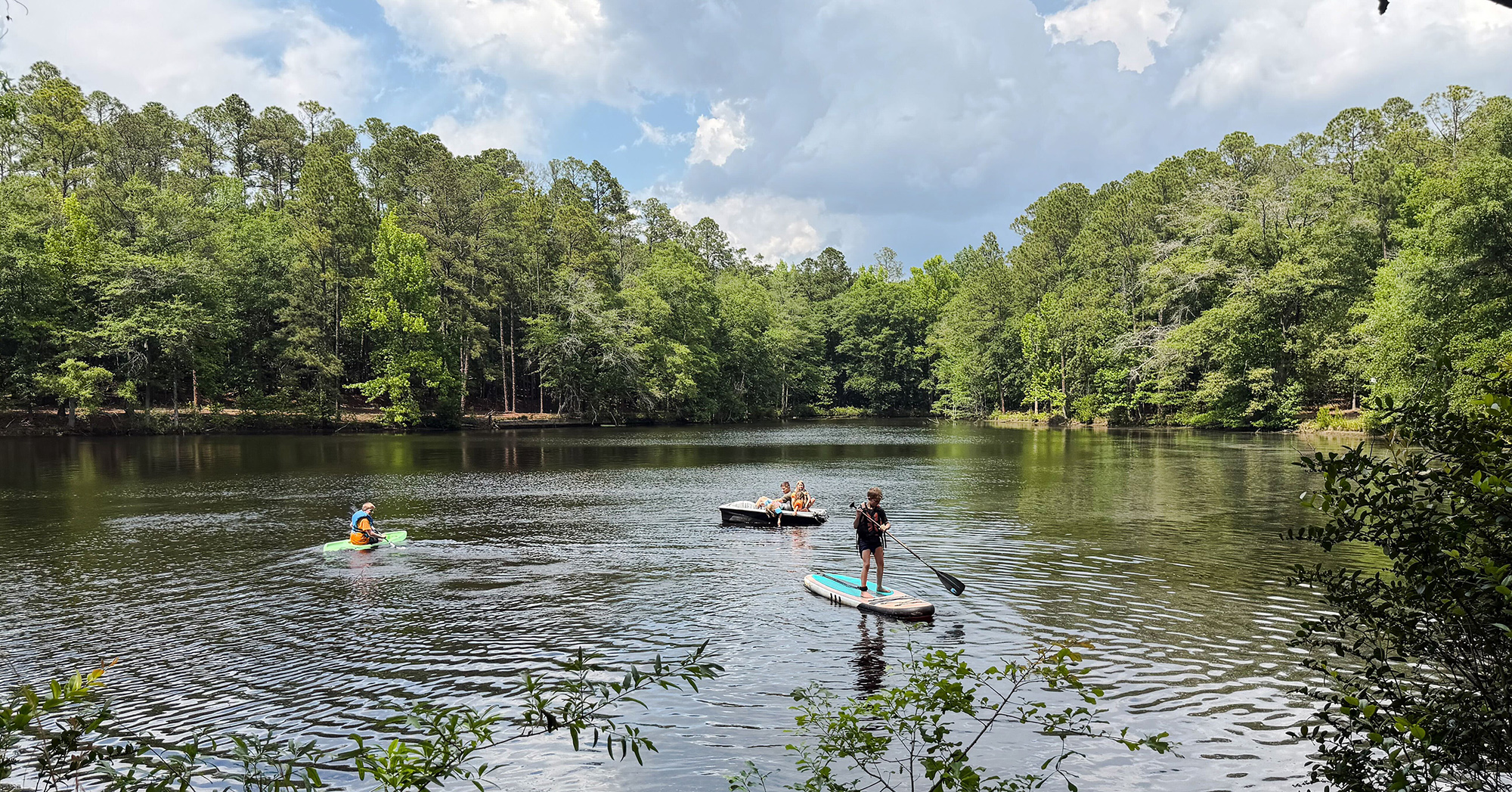 Two people paddleboarding on a calm lake near a forested shoreline, with a small boat carrying several riders nearby under a partly cloudy sky.