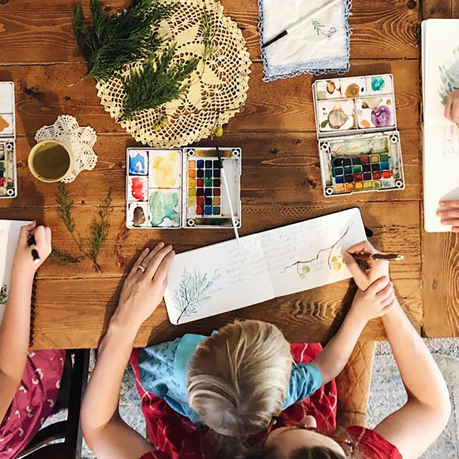 Overhead view of people painting watercolors at a wooden table with paint palettes, brushes, and a greenery centerpiece on a doily.