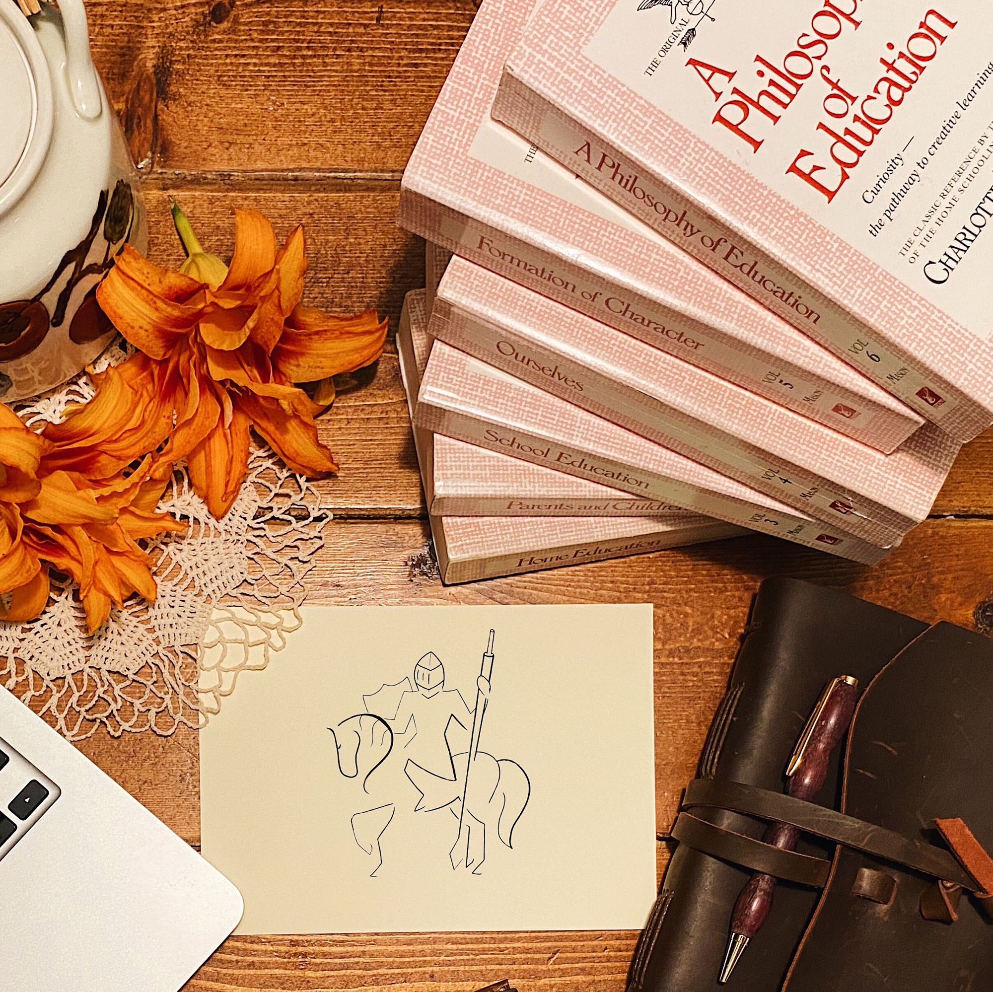 Top-down view of a wooden desk with orange flowers, a lace doily, a white laptop, and a stack of pink Philosophy of Education textbooks.