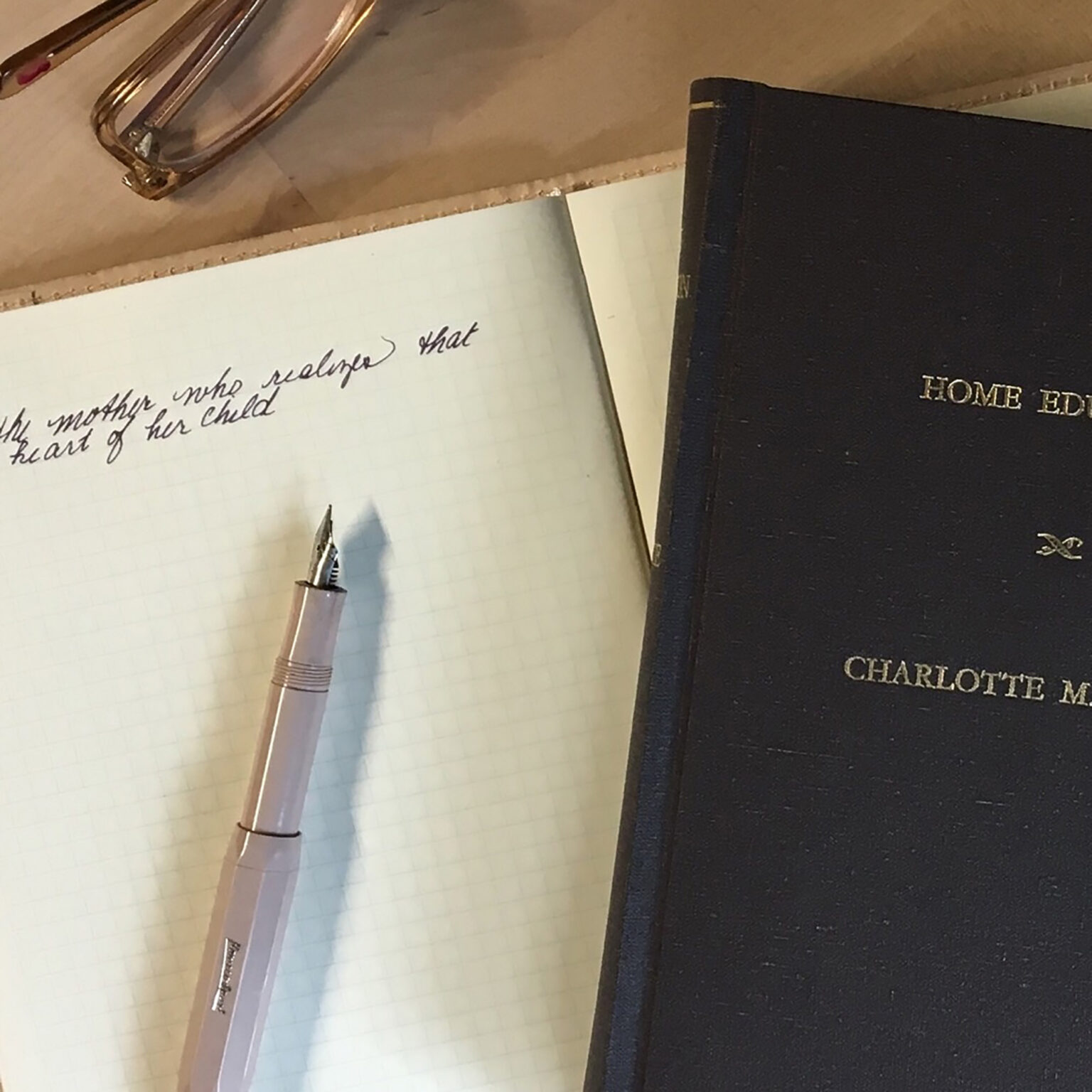 Close-up of a desk with a handwritten page, a glass fountain pen, and a dark notebook labeled 'HOME EDU' on a wooden surface.
