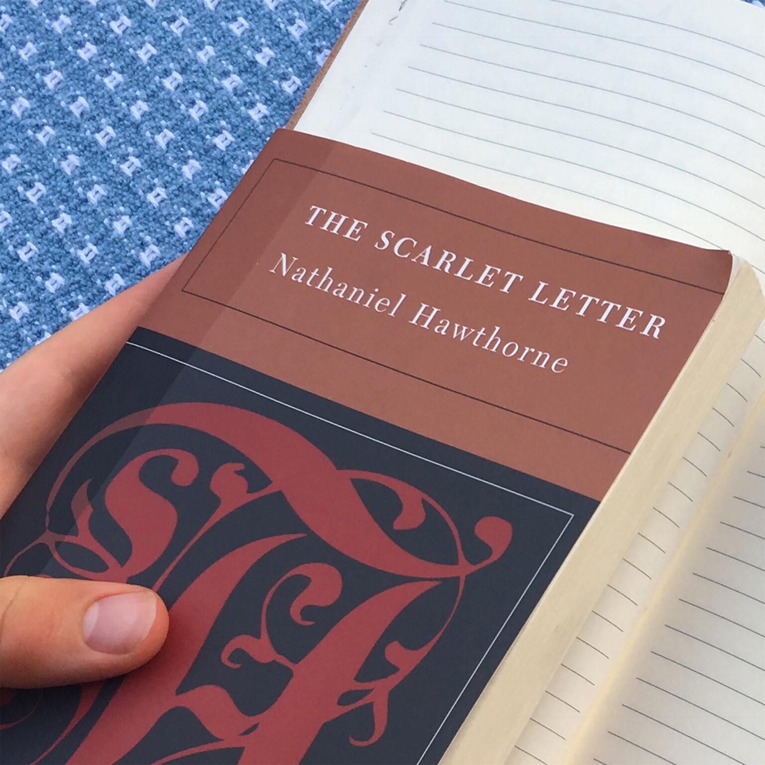 Close-up of a hand holding a brown paperback titled 'The Scarlet Letter' by Nathaniel Hawthorne, with a blue patterned carpet and a lined notebook in the background.