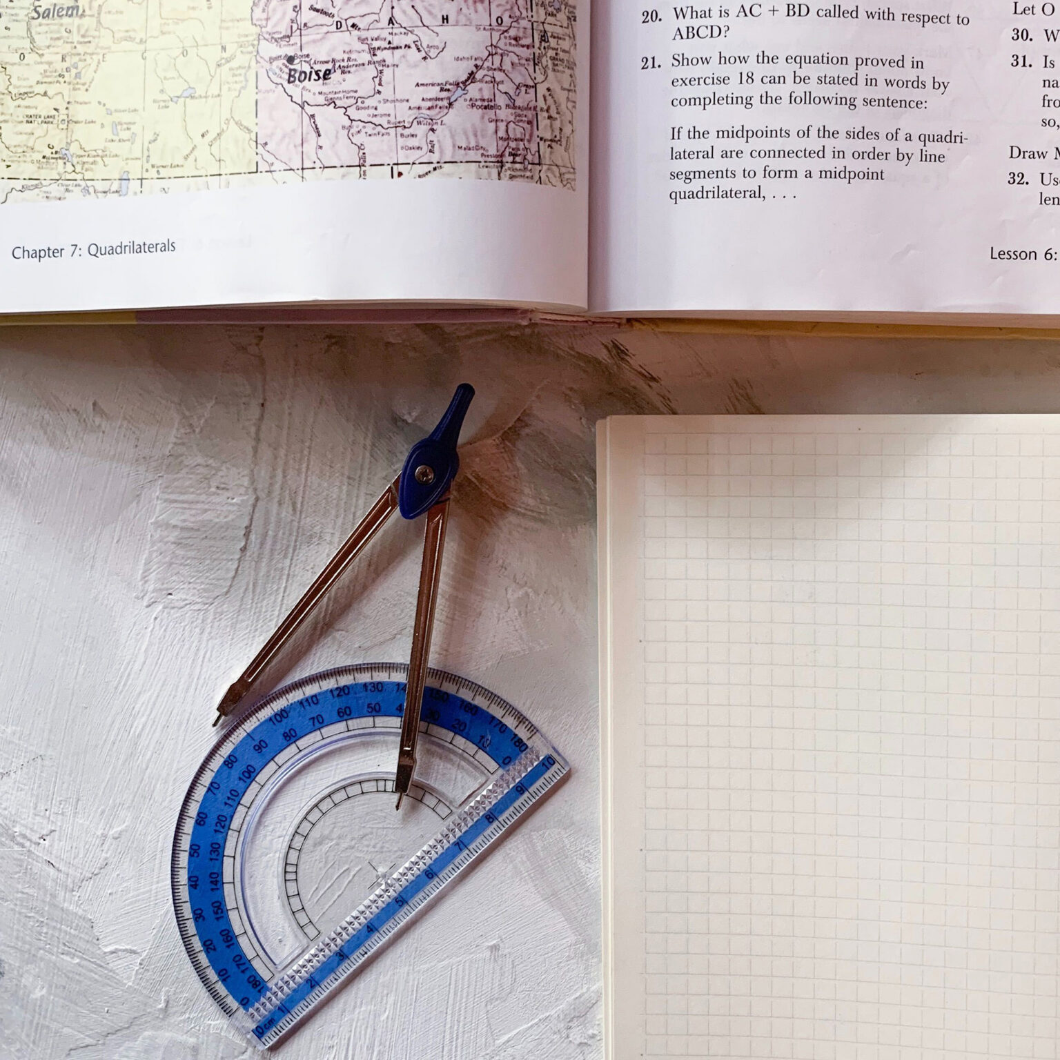 Protractor, compass, and ruler laid on a desk next to a grid notebook and map page.