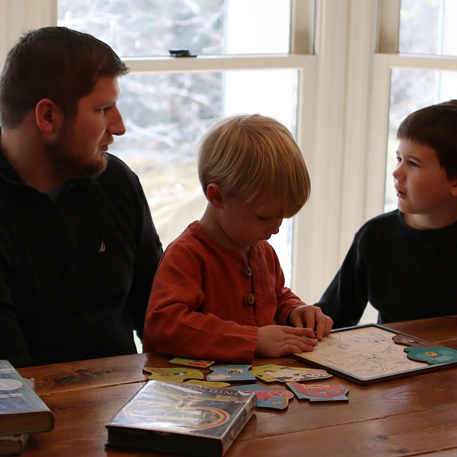 Man and two children seated at a wooden table, playing a board game with colorful puzzle pieces as daylight comes through a window behind them.