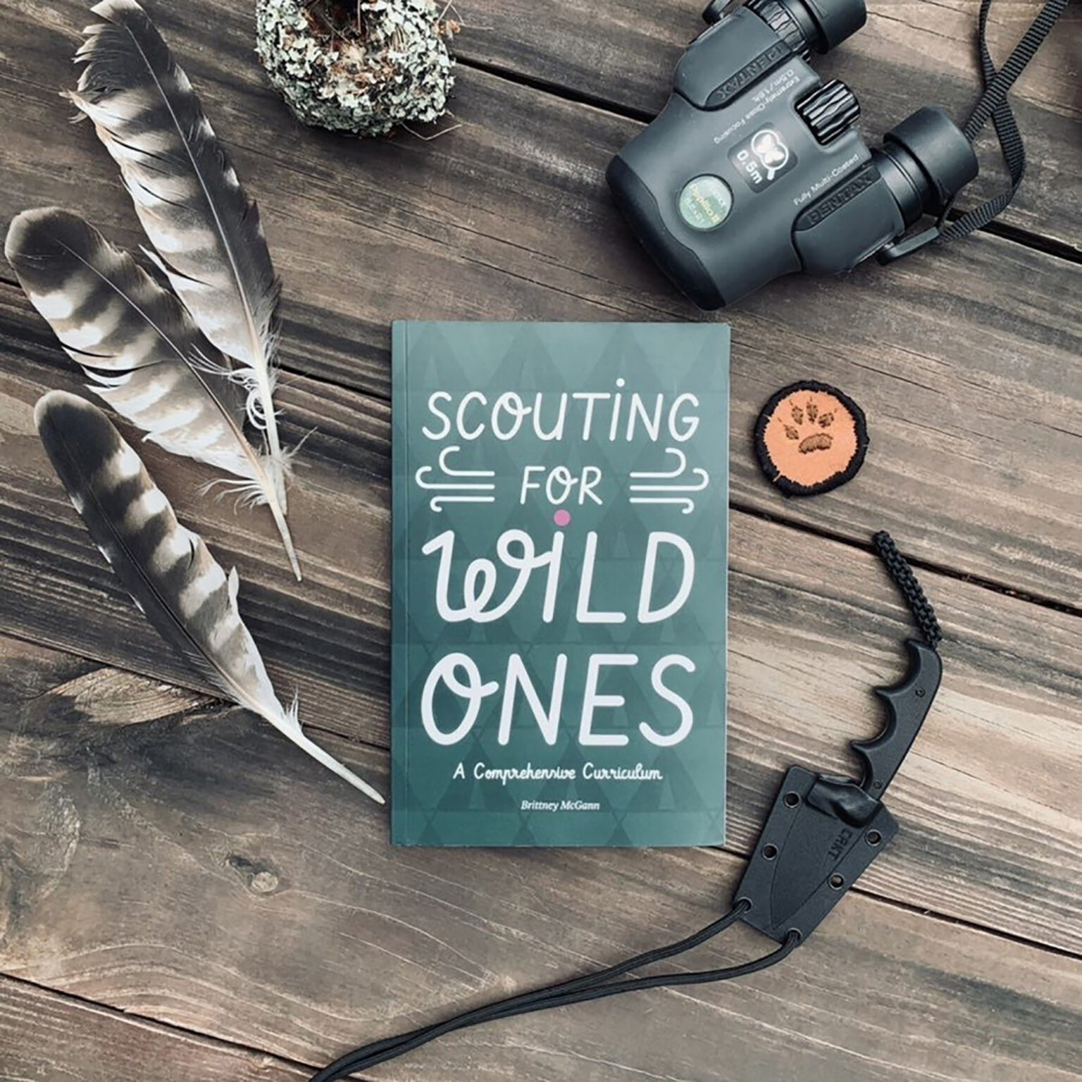 Notebook titled 'Scouting for Leads' with a compass motif sits on a wooden table beside binoculars, a feather, and a pine cone pieces nearby.