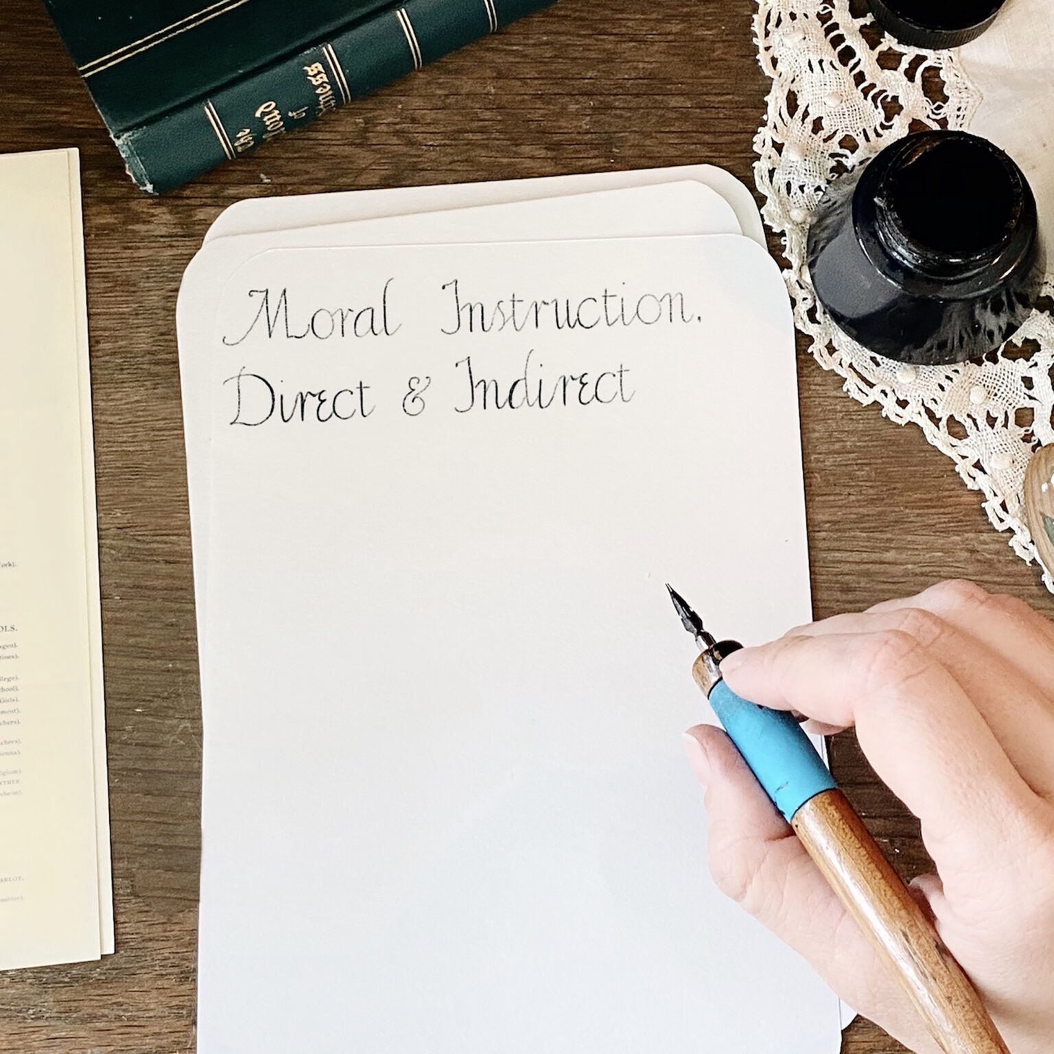 Hand with a blue fountain pen over a notebook page reading 'Moral Instruction Direct & Indirect' on a wooden desk with an ink bottle nearby.
