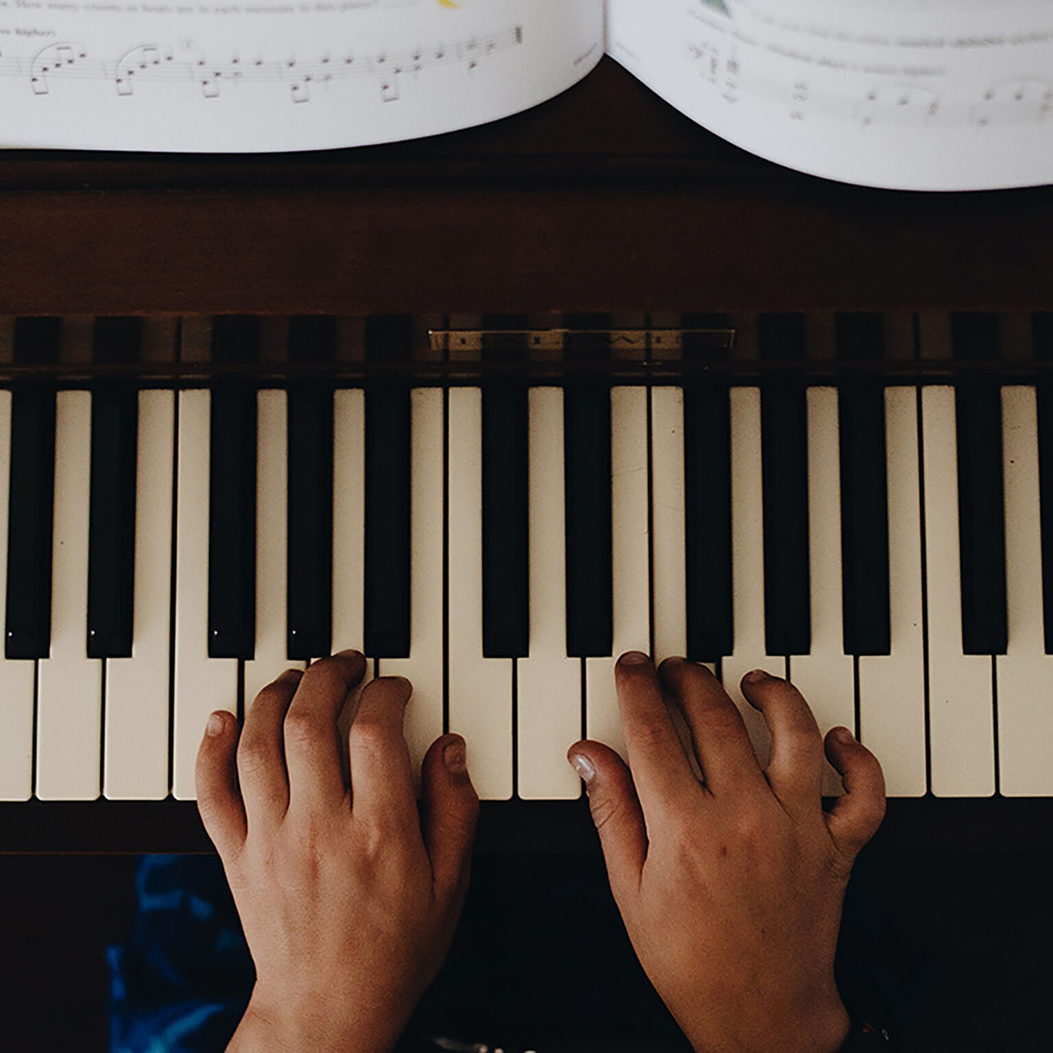 Top-down view of hands playing a piano keyboard with sheet music at the top edge of the frame.