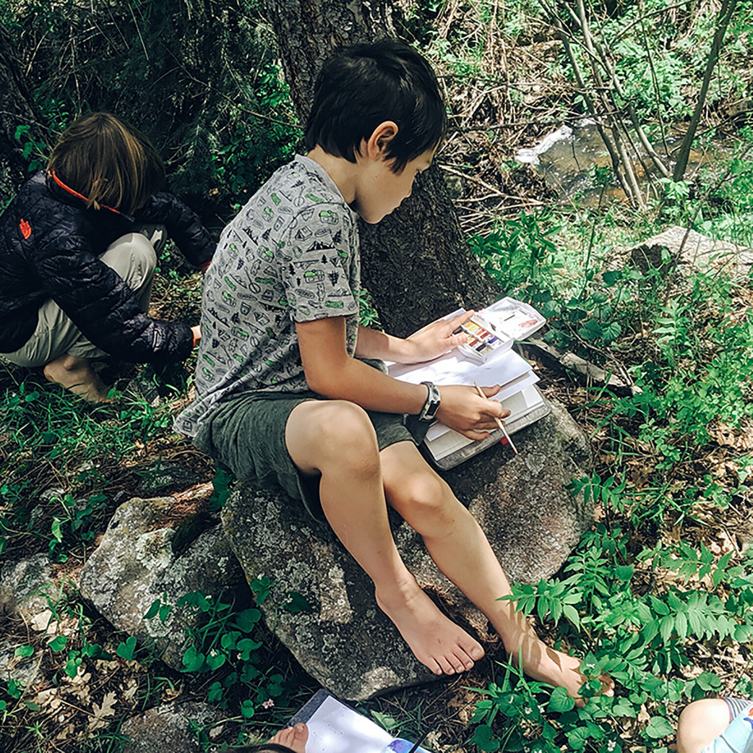 Two children sit on a rocky ground in a forest, looking at/maps spread out on the ground amid greenery.