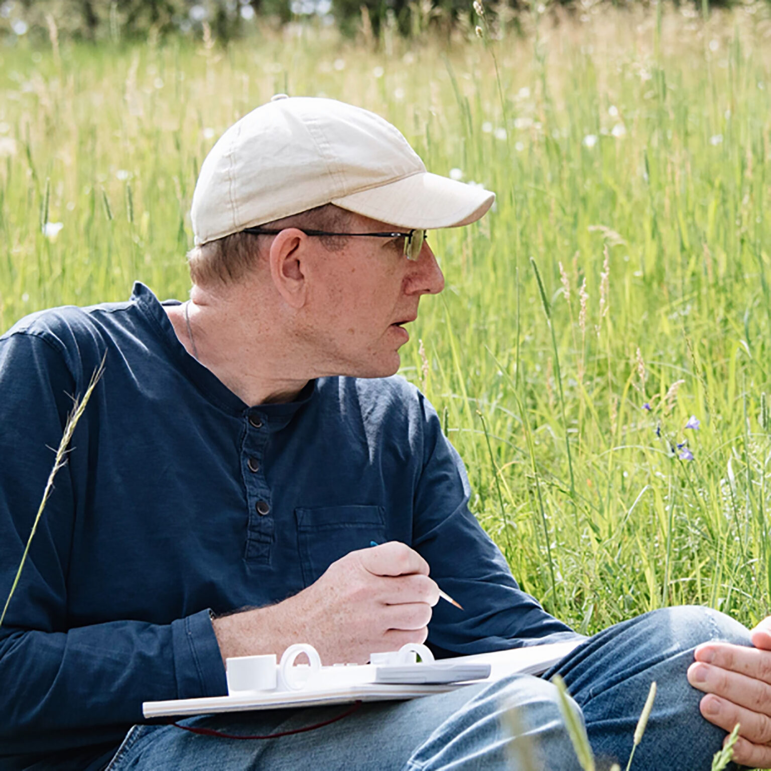 Man in a beige cap and dark shirt sits in a grassy field, assembling a white plastic model kit on his lap.