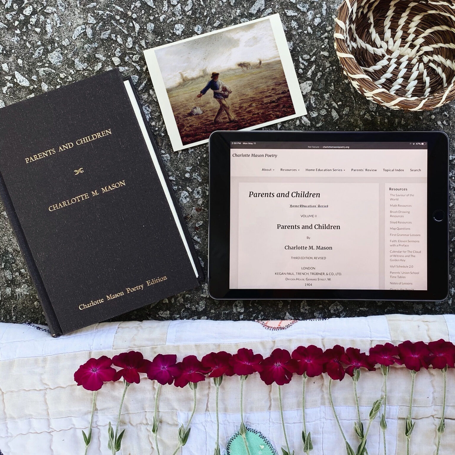 Overhead scene with a dark hardcover poetry book, a small landscape painting postcard, a woven basket, and a tablet displaying a 'Parents and Children' page.
