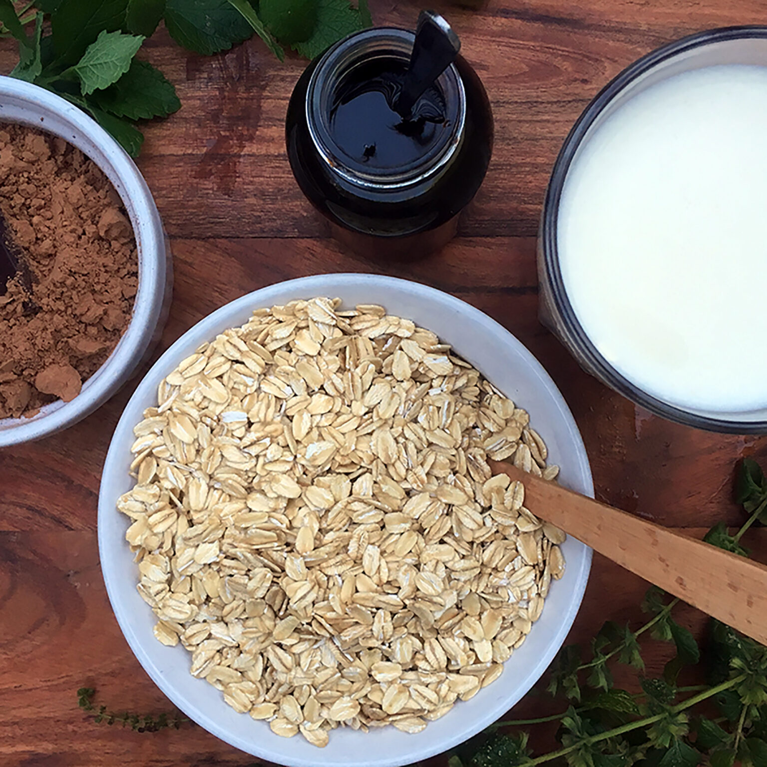 Overhead view of a white bowl filled with oats on a wooden table, with a jar of dark syrup and green leaves nearby.